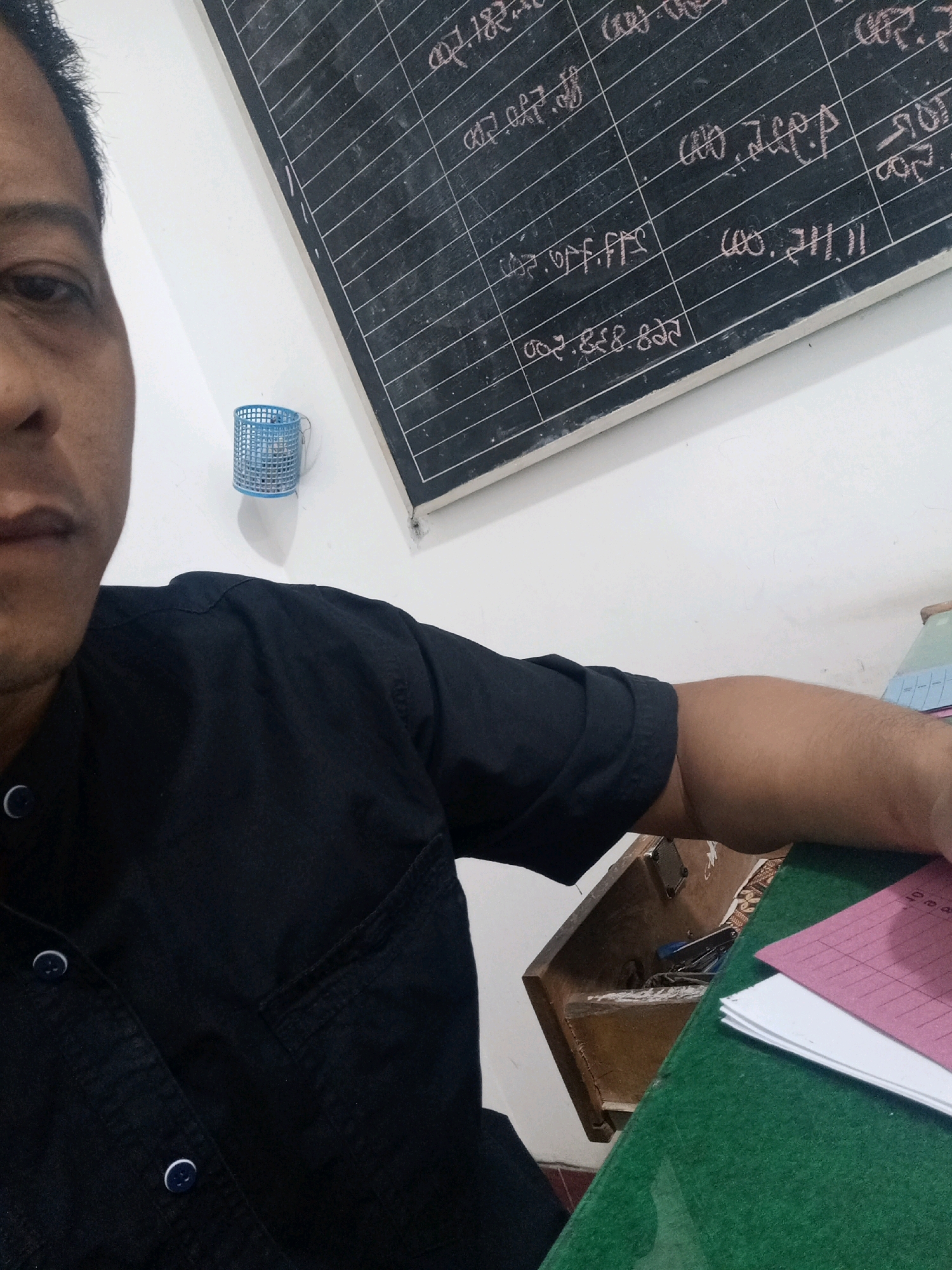 A man's face and torso are partially visible on the left side of the image. He is wearing a black collared shirt with buttons. Behind him, a blackboard with rows of handwritten notes is mounted on a white wall. A small blue plastic basket is attached to the wall below the blackboard. To the right of the blackboard, a wooden desk with a green surface is visible. Several papers are stacked on the desk, one of which is pink and has a grid pattern. The context suggests this is a classroom or office setting in Begajah, Indonesia.