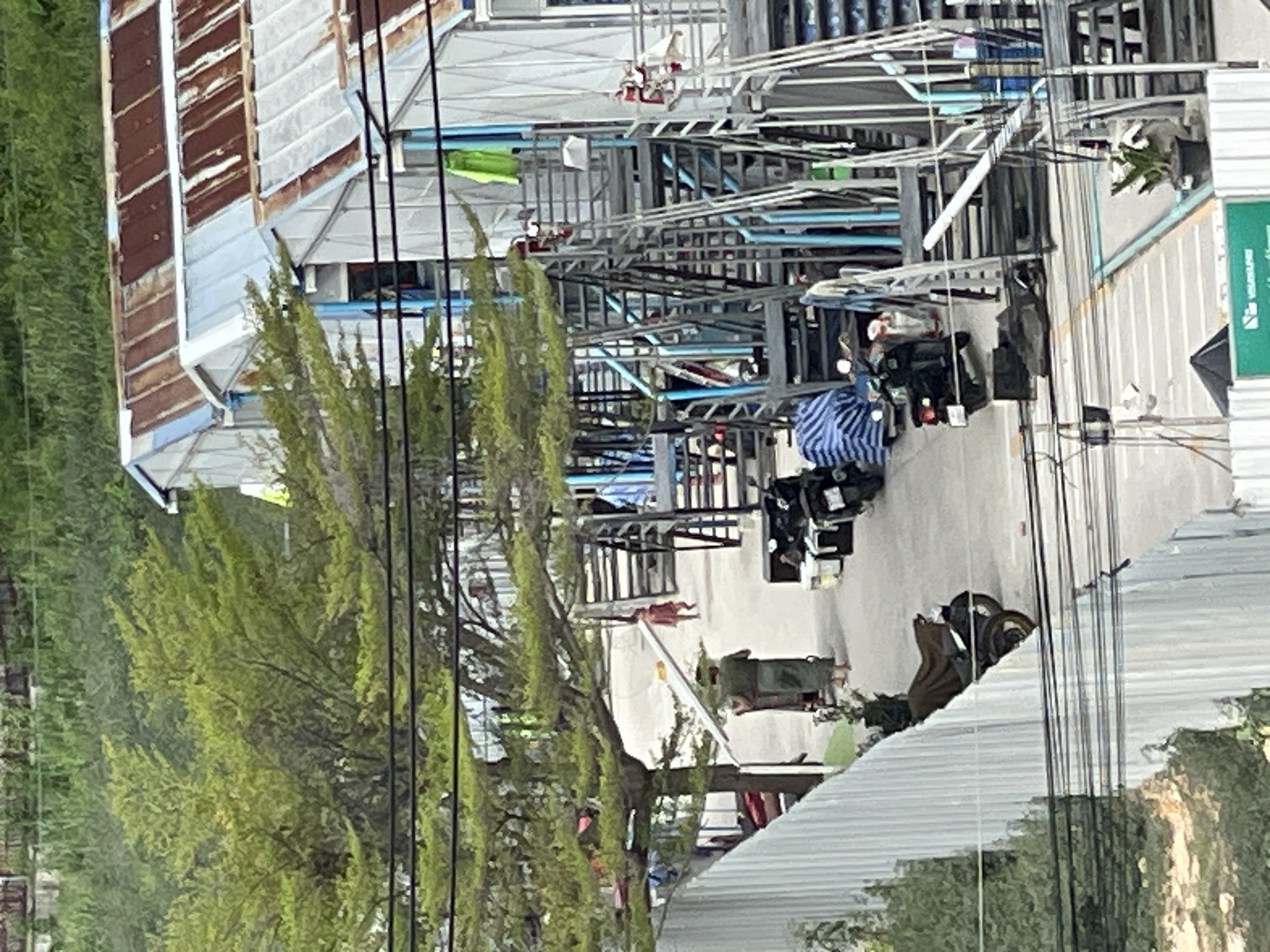 The image depicts a scene in Pattaya City, Thailand, focusing on an outdoor area adjacent to a building with a corrugated metal roof and white siding. Several external staircases, constructed with gray metal and blue pipes, lead up to this building. In the foreground, there is a white corrugated metal fence, and beyond it, a paved walkway or road.
Several motorcycles are parked along the walkway. One person, dressed in a dark top and a light-colored sarong or skirt, stands near a cart, appearing to be engaged in some activity. Further down the walkway, a smaller figure, likely a child, is visible. There are also potted plants and various items placed around the area, including what appear to be storage containers. Laundry, including a bright green garment, is hanging on a line or draped over structures near the building. The overall environment suggests a residential or communal living area, possibly with a commercial aspect given the presence of shops or stalls implied by the building's design. Overhead power lines are visible across the top of the image, indicating an urban setting.