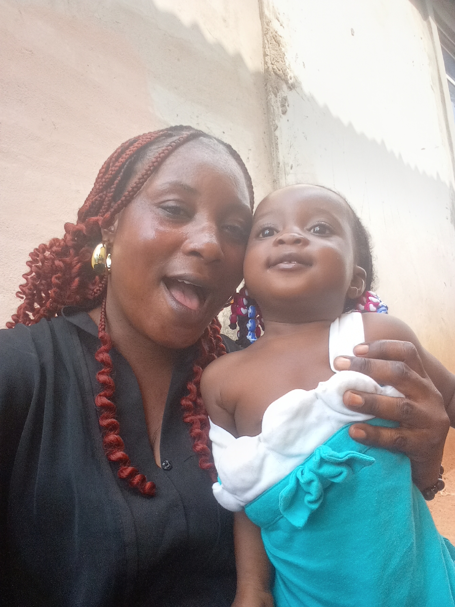 The image shows a woman and a child in Doma, Nigeria. The woman, with red braided hair and gold hoop earrings, is wearing a black shirt and has her mouth open. She is holding a baby dressed in a light blue outfit with a white top. The baby is looking upwards with a slight smile. The background is a light-colored wall with some discoloration.