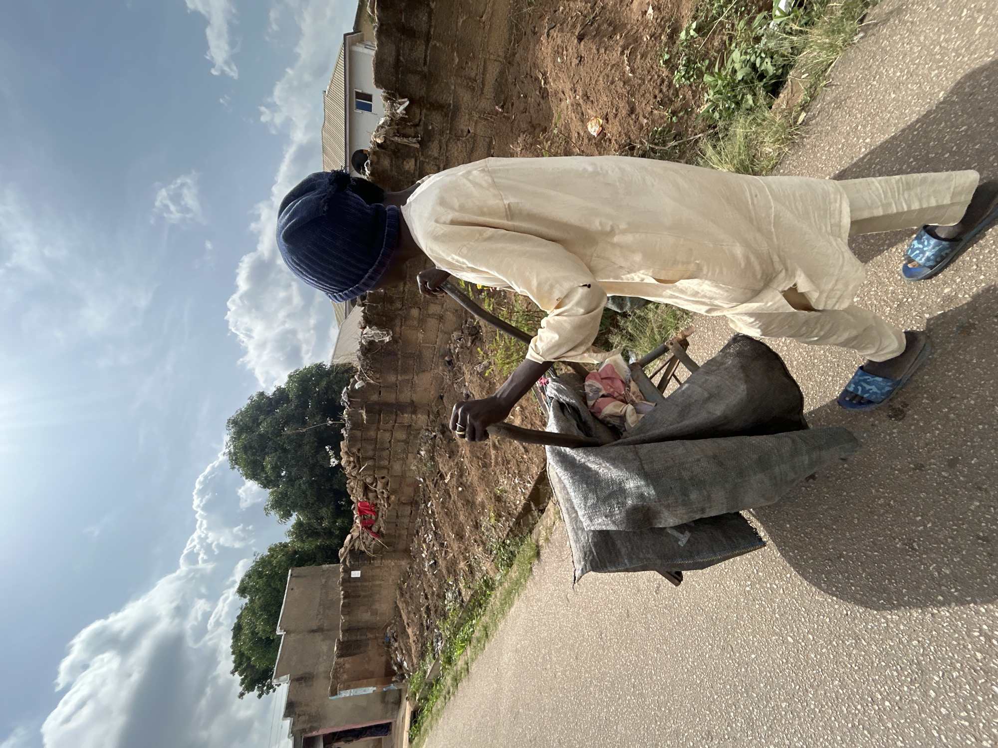 A person in a light-colored tunic, dark blue cap, and blue sandals is pulling a cart covered in a gray tarp down an asphalt road in Bar Jirgi Summa, Nigeria. The cart appears to contain items wrapped in the tarp, with a glimpse of pink cloth visible. The person's dark skin contrasts with the tunic. To the left, a brick wall with debris accumulated at its base runs alongside the road, with a few small green plants growing nearby. A building is partially visible behind the wall. The sky is light blue with white clouds.
