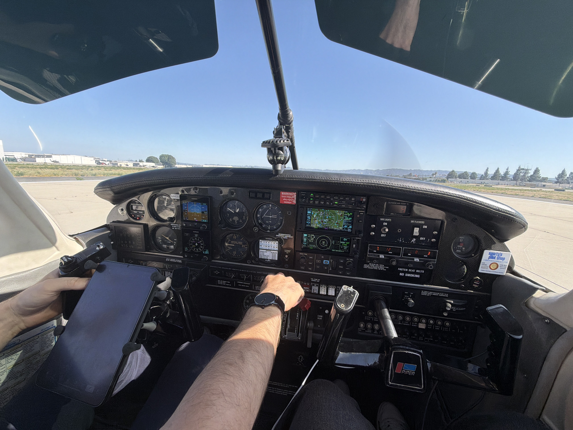 The image captures the interior of a Piper aircraft cockpit, on the ground. The pilot, wearing a watch, is operating the aircraft's controls. The dashboard is visible, with various instruments and switches, including the navigation lights, sky flashers, and carb heat. A tablet is mounted on a holder. Outside the cockpit window is an airfield runway. The plane is on the ground near the runway, which is a concrete slab. Visible outside the window are buildings that look like warehouses and some grass and trees. The aircraft identifier is visible as "N4409J". The location seems to be near a city but hard to identify it by looking the image. The location in Los Angeles, United States of America is impossible to determine without further information.