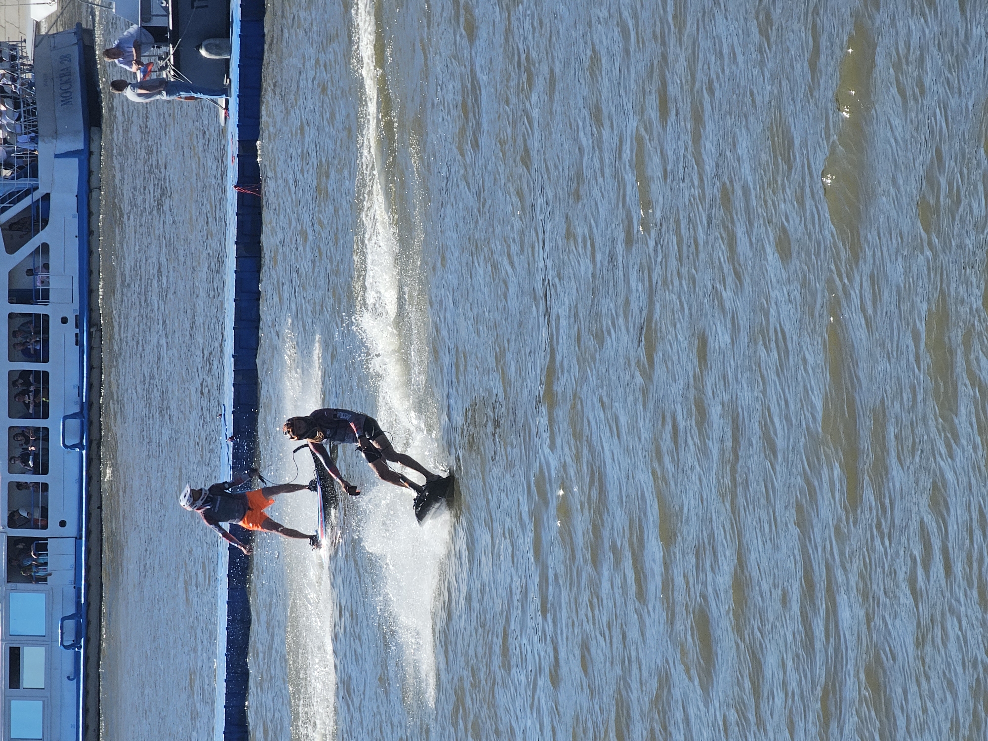 The image shows a body of water with several elements related to water sports and a boat. In the foreground, a person is actively engaged in wakeboarding, creating a significant splash. The wakeboarder is wearing a helmet and athletic clothing. Behind them, another individual is also on a wakeboard, appearing to be either waiting or also participating in the activity. In the background, a large white boat with blue accents is visible, with people on its upper deck. The boat has the word "МОСКВА 28" written on its side. A blue floating platform is situated in the water, with two individuals standing on it, seemingly observing the water sports or preparing for their turn. The environment is sunny, with clear blue water and a bright sky. The scene appears to be taking place on a river or a large lake within a city, as suggested by the presence of the boat and the urban backdrop implied by the boat's name, which suggests Moscow.