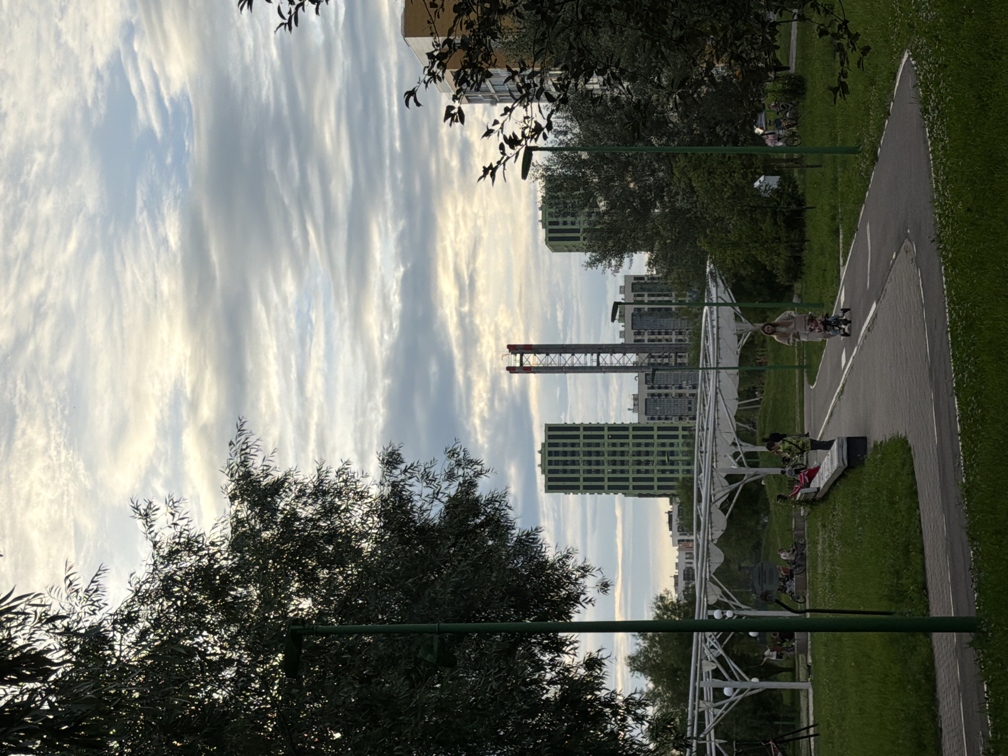 This image depicts a contemporary urban park scene in Kazan, Russia, likely in a newly developed residential area. In the background, modern high-rise buildings, some with distinctive green facades, punctuate the skyline under a cloudy sky. A prominent red and white tower structure is also visible.
In the foreground and mid-ground, the park features paved pathways for walking and cycling, with a woman and child on a tricycle moving along one. Green grass areas with scattered white flowers are abundant. Several people are visible sitting on a bench and gathered in a shaded area under a white, arched canopy structure, some with bicycles. Large trees, silhouetted against the sky, frame the scene, with streetlights visible on green poles. The overall atmosphere suggests a relaxed, communal outdoor space designed for recreation and leisure in a modern urban setting.