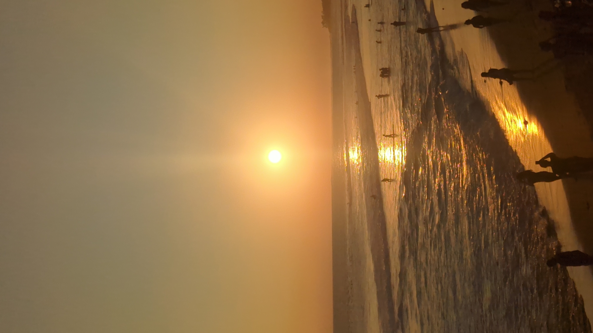 The image depicts a beach at sunset in North Kuta, Indonesia. The sun is positioned near the horizon, casting a bright glow on the water. Silhouettes of people are visible along the shoreline, some standing on the sand and others wading in the water. The waves reflect the sunlight, creating a shimmering effect. The sky is a gradient of orange and yellow hues.