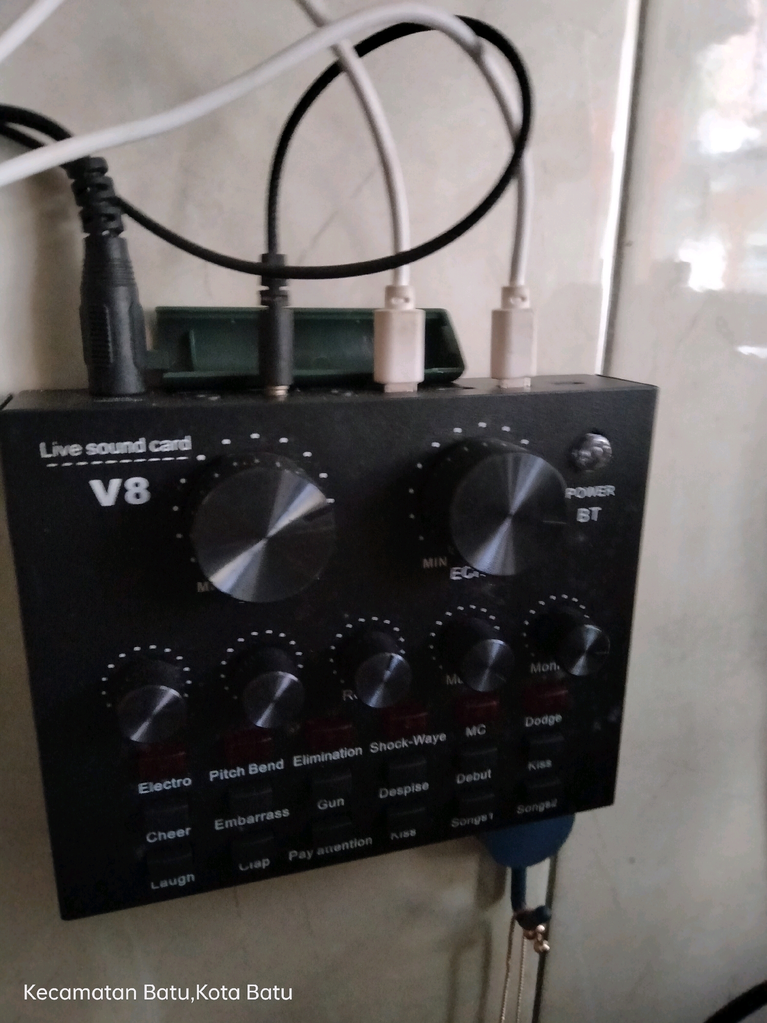The image shows a black V8 live sound card with multiple knobs and buttons on its front panel. Several cables are plugged into the top of the device, including a black cable with a coiled connector, and two white cables with USB connectors. The sound card is positioned in front of a textured, light-colored wall. Below the sound card, a white text overlay reads "Kecamatan Batu, Kota Batu", indicating the location as Batu City, Indonesia. The overall lighting suggests an indoor setting.