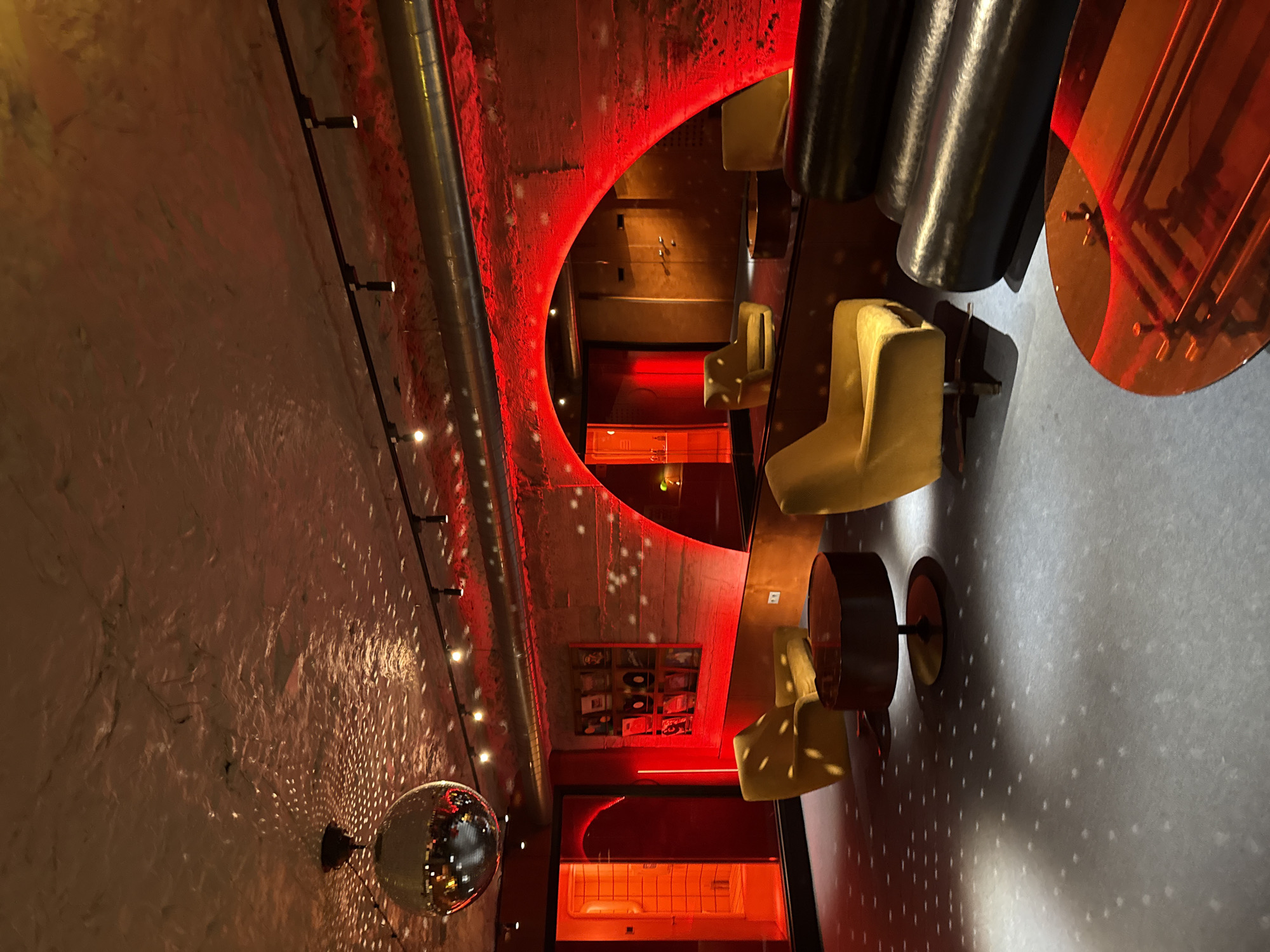 This image captures a vibrant, modern interior space, likely a bar or lounge, characterized by a striking red lighting scheme. The photograph is oriented vertically, looking across a room with high ceilings, suggesting a dynamic and trendy establishment in Yerevan, Armenia.
On the left, a large, silver disco ball is affixed to a textured, light-colored wall, casting numerous small, sparkling reflections across its rough surface. Above this, a dark rail with several small, bright spotlights runs horizontally along the wall, further illuminating the area. Exposed metal pipes, possibly ventilation or structural elements, run along the upper edge of this wall and into the ceiling, adding an industrial aesthetic.
The central feature is a large, crescent-shaped opening or mirror outlined by intense red light, which acts as a dramatic focal point. This mirror reflects a deeper section of the venue, revealing a wall-mounted display of what appear to be vinyl records or artworks, and a further red-lit corridor or room with a white fixture visible in the background.
In the foreground and partially reflected, several contemporary armchairs upholstered in a light yellow or mustard fabric are arranged with small, dark, round tables, creating intimate seating areas. On the right side of the frame, large, dark cylindrical objects hang from the ceiling, alongside a semi-transparent, reddish-orange circular panel, contributing to the eclectic decor. The floor appears to be a smooth, dark surface, reflecting some of the overhead light.
The overall mood is energetic and sophisticated, illuminated primarily by artificial light, suggesting nighttime. No people are visible, but the setup implies a setting for social gatherings or entertainment. There is no discernible text in the image.