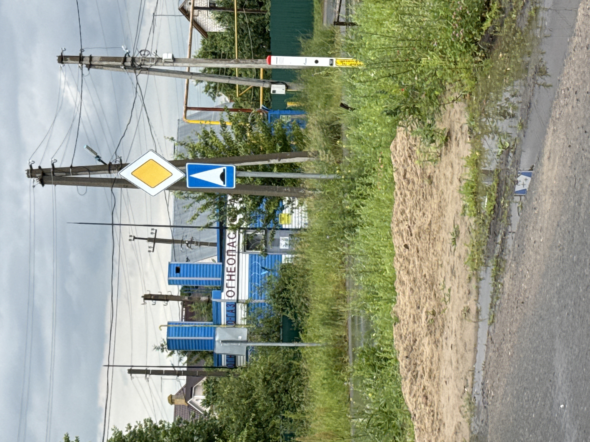 The image depicts an outdoor scene, likely in a rural or suburban area, with a focus on infrastructure and vegetation. Several utility poles with numerous wires dominate the left side of the frame, suggesting a power or communication lines. Next to these poles are traffic signs, including a yellow diamond-shaped sign and a blue rectangular sign with a white arrow indicating a turn.
In the background, there's a building with blue paneling and white trim, possibly a commercial establishment or an industrial facility. The word "ОГНЕОПАС" is partially visible in white lettering on the blue facade, which translates to "FLAMMABLE" in Russian, indicating the nature of the goods or services offered. There are also smaller structures, possibly storage units or sheds, also clad in blue. The surrounding area is filled with lush green vegetation, including tall grass and bushes, with some trees visible. A dirt mound or pile of material is prominent on the right side of the image, adjacent to an asphalt road that appears wet, suggesting recent rain or a puddle. The sky is overcast with gray clouds, implying a cloudy or potentially rainy day.
There are no people visible in the image. The overall impression is one of a functional, perhaps industrial, roadside area. The time of day is difficult to ascertain precisely due to the overcast sky, but it appears to be daytime. The location cues, such as the Cyrillic text on the building, and the general landscape, are consistent with a setting in Russia.
