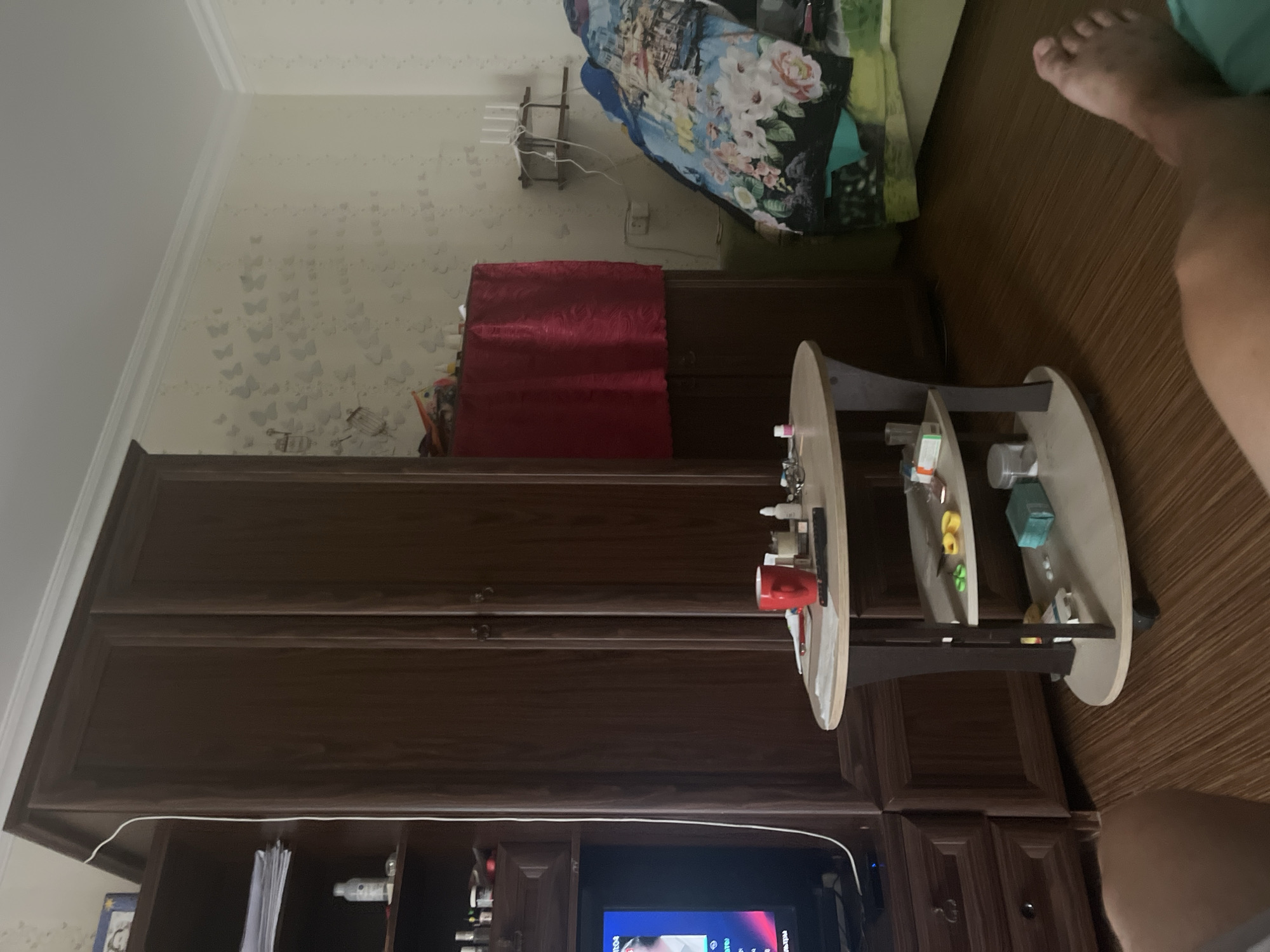 The image depicts a domestic interior scene, likely a bedroom or living room, with a focus on furniture and personal items. Prominently featured is a large, dark wood wardrobe. To the left of the wardrobe is a smaller, dark wood shelving unit with a television set. On top of the shelving unit are stacks of papers and a small bottle.
In front of the wardrobe, a small, oval-shaped table with two tiers holds various objects, including a red mug, a remote control, a bottle, glasses, and what appear to be medication boxes. The lower shelf of this table contains smaller items like yellow and green objects, a green rectangular object, and small white pills.
To the right of the wardrobe, a bed is visible, covered with a comforter featuring a floral and ship motif. A green piece of fabric is partially visible beneath the comforter.
The walls appear to be a light color with a subtle patterned wallpaper. A small shelf with a white router and wires is mounted on the wall above the bed. The flooring is a wooden laminate. A human foot, partially in frame at the bottom right of the image, suggests a person is present in the room, possibly lying on the bed or floor. The overall impression is one of a lived-in space with everyday objects.