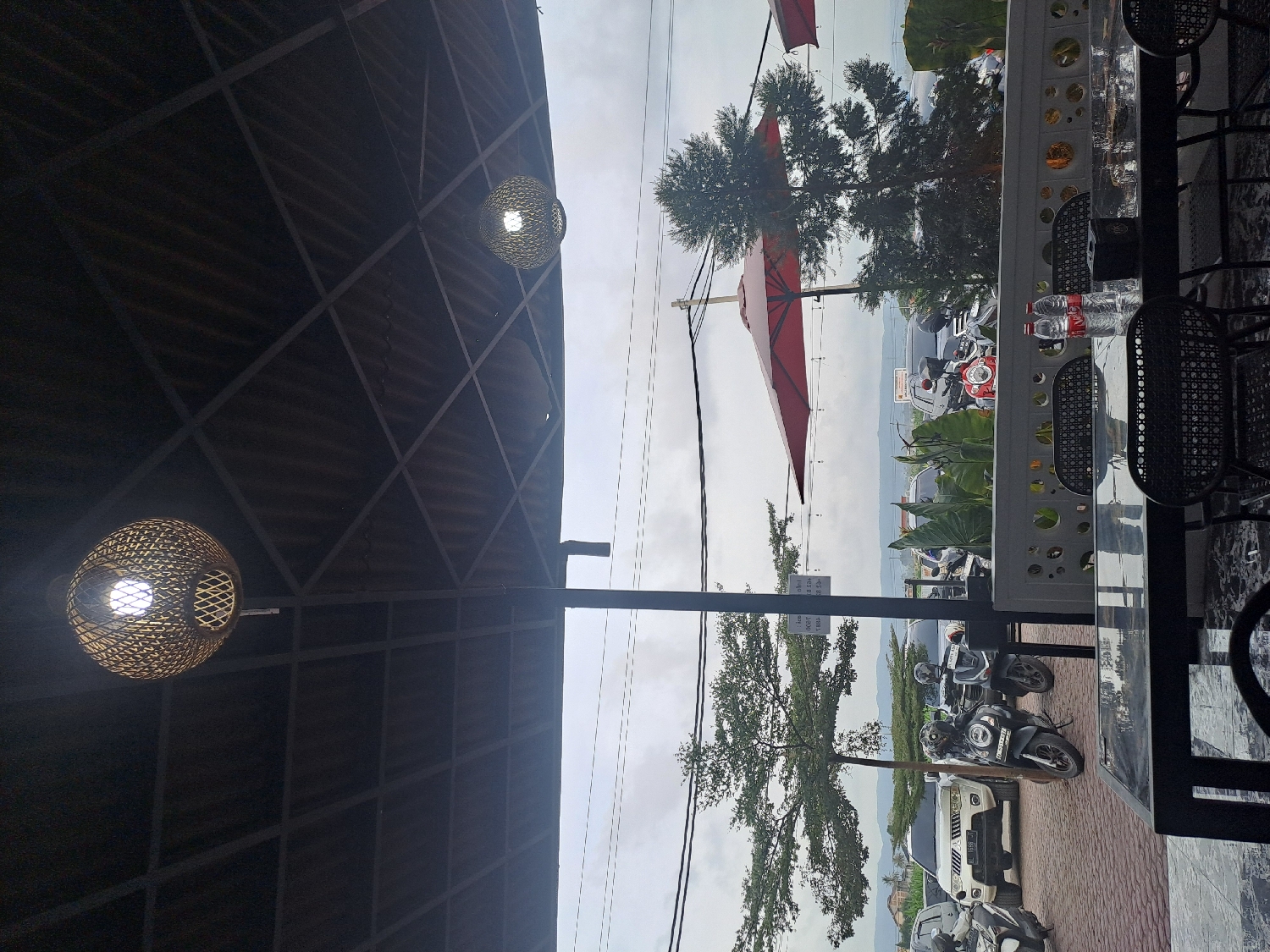 This image captures a semi-outdoor seating area, likely part of a cafe or restaurant, offering a view of a street and distant hills. In the foreground, two spherical light fixtures with woven designs hang from a dark, industrial-style ceiling with a grid pattern. The lights within these fixtures are illuminated.
To the right, a dark table with a decorative, patterned surface is visible, along with two chairs featuring woven backs and seats. Two plastic water bottles are placed on the table. A white railing or barrier with circular cutouts separates the seating area from the exterior.
Beyond the railing, a street or parking area is visible, populated with several parked motorcycles and at least one car. A large, red umbrella or canopy partially extends into the frame from the right. Green trees and other foliage line the street. In the background, rolling hills or mountains are seen under an overcast sky. A small white sign near the street has partially legible text, including "dilarang parkir" (Indonesian for "no parking").
The scene appears to be set during the daytime under cloudy conditions. The overall atmosphere suggests a relaxed, casual dining or waiting area in Glameunasah Baro, Indonesia. No people are visible in the frame.