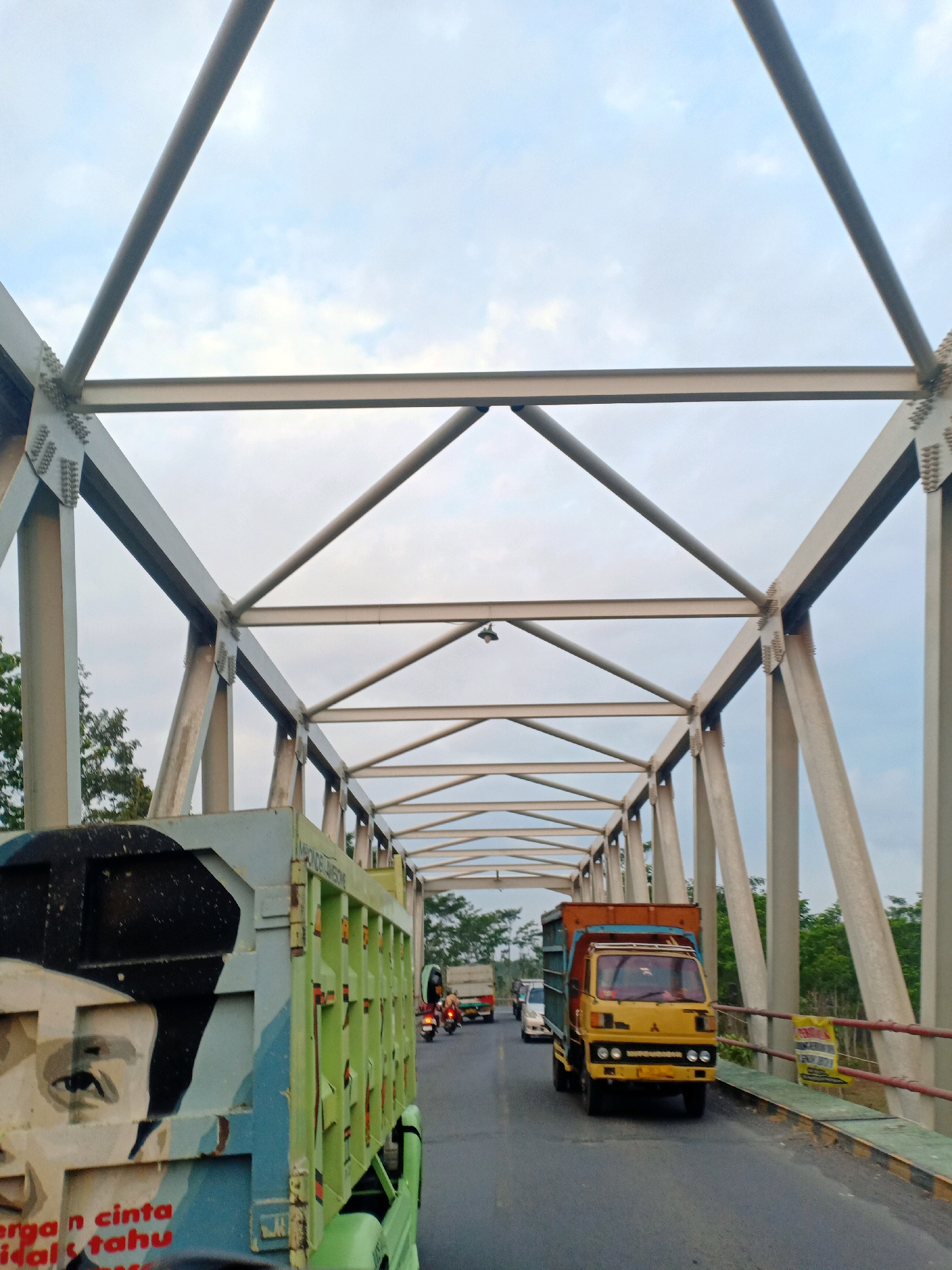 This is a low-angle, outdoor, eye-level shot taken on a bridge. The bridge has a steel truss structure overhead. The road is asphalt, and there are vehicles driving on it, including a green truck with a large, abstract face painted on its side, a yellow truck, and a motorcycle. The sky is overcast with some clouds. The scene appears to be in a rural or suburban area, with trees visible in the background.