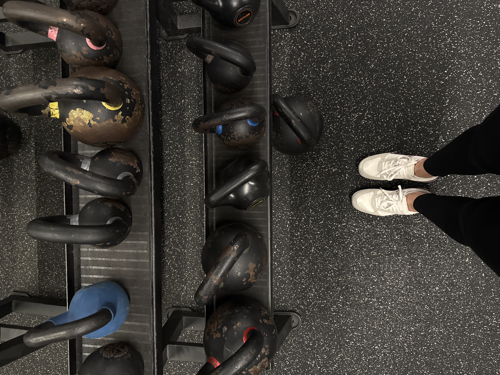 The image shows a gym setting with a rack of kettlebells and a person's feet wearing white sneakers and black leggings. The kettlebells are of various sizes and weights, some showing signs of wear and tear, with chipped paint revealing rust underneath. One kettlebell is partially covered in blue material. The floor is a black, speckled rubber matting common in gyms. The lighting suggests an indoor setting, possibly during the day given the quality of light. The overall atmosphere is functional and focused on exercise equipment. No specific time of day or weather conditions are discernible. There is no visible text in the image apart from a small "KETTLE" logo on one of the kettlebells. The scene is set in a gym in Sochi, Russia.