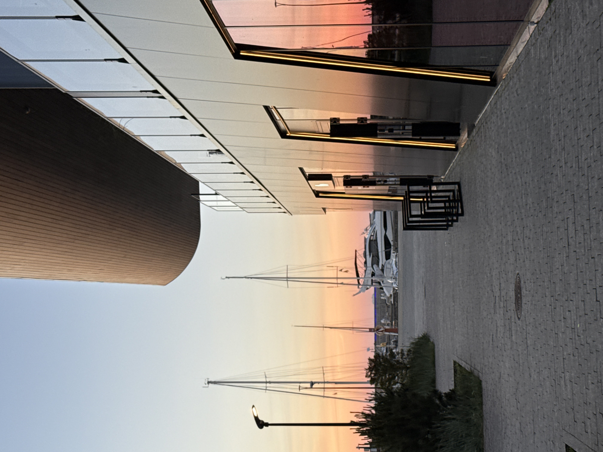 The image captures a marina scene at dusk in Tallinn, Estonia. The foreground features a paved walkway made of grey interlocking stones, leading towards a modern building with a sleek, dark facade and large glass windows reflecting the warm, orange and pink hues of the sunset. Vertical yellow lights accent the windows and doorways of the building. To the left of the walkway, there is a patch of green grass and dark green bushes.
Beyond the walkway and building, several sailboats with tall masts are anchored in the marina. One prominent white yacht is docked closer to the shore. The sky is a gradient of pale blue at the top, transitioning to vibrant orange and pink as it nears the horizon, indicating the setting sun. A streetlamp with a single light fixture stands on a black pole to the left of the scene, near the bushes. The overall atmosphere is tranquil and serene, highlighting the juxtaposition of modern architecture with the natural beauty of a marina at sunset.