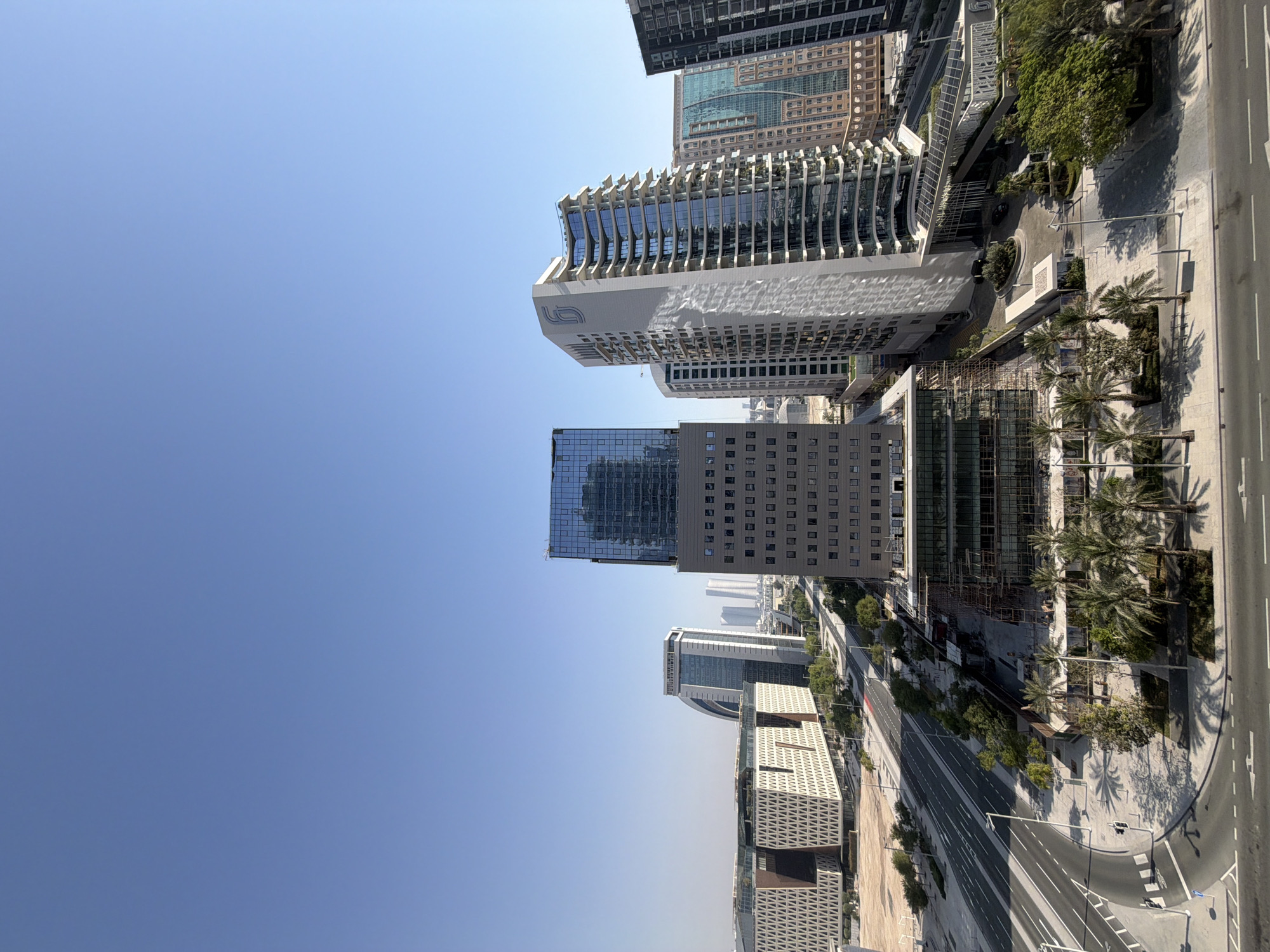 This image captures a cityscape under a clear blue sky. The scene is dominated by modern architecture, featuring a variety of tall buildings with glass facades and intricate designs. In the foreground, a multi-lane road curves through the urban landscape, with streetlights and palm trees lining the sidewalks. Construction is visible on one of the buildings, indicated by scaffolding. The overall impression is one of a bustling, contemporary city.