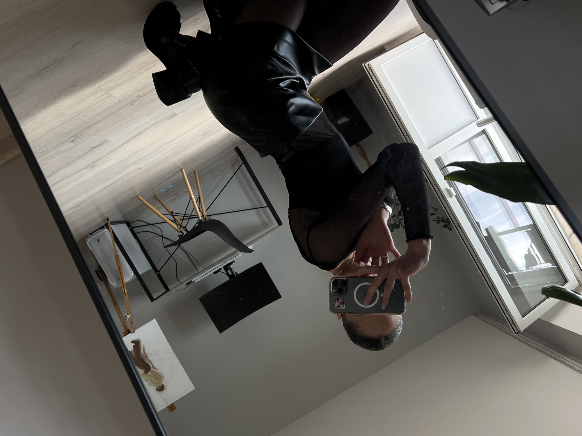 A person, seen from the reflection in a mirror, is kneeling on a light-colored wooden floor. The person is wearing a black sheer long-sleeved top and a black leather skirt. They are holding a smartphone up to take a selfie. The background shows a room with a desk, a computer monitor, a painting on an easel, and a large window with white doors. The scene is indoors, likely in a residential setting. There is no information in the image to indicate the location is in the city of Ternopil, Ukraine.