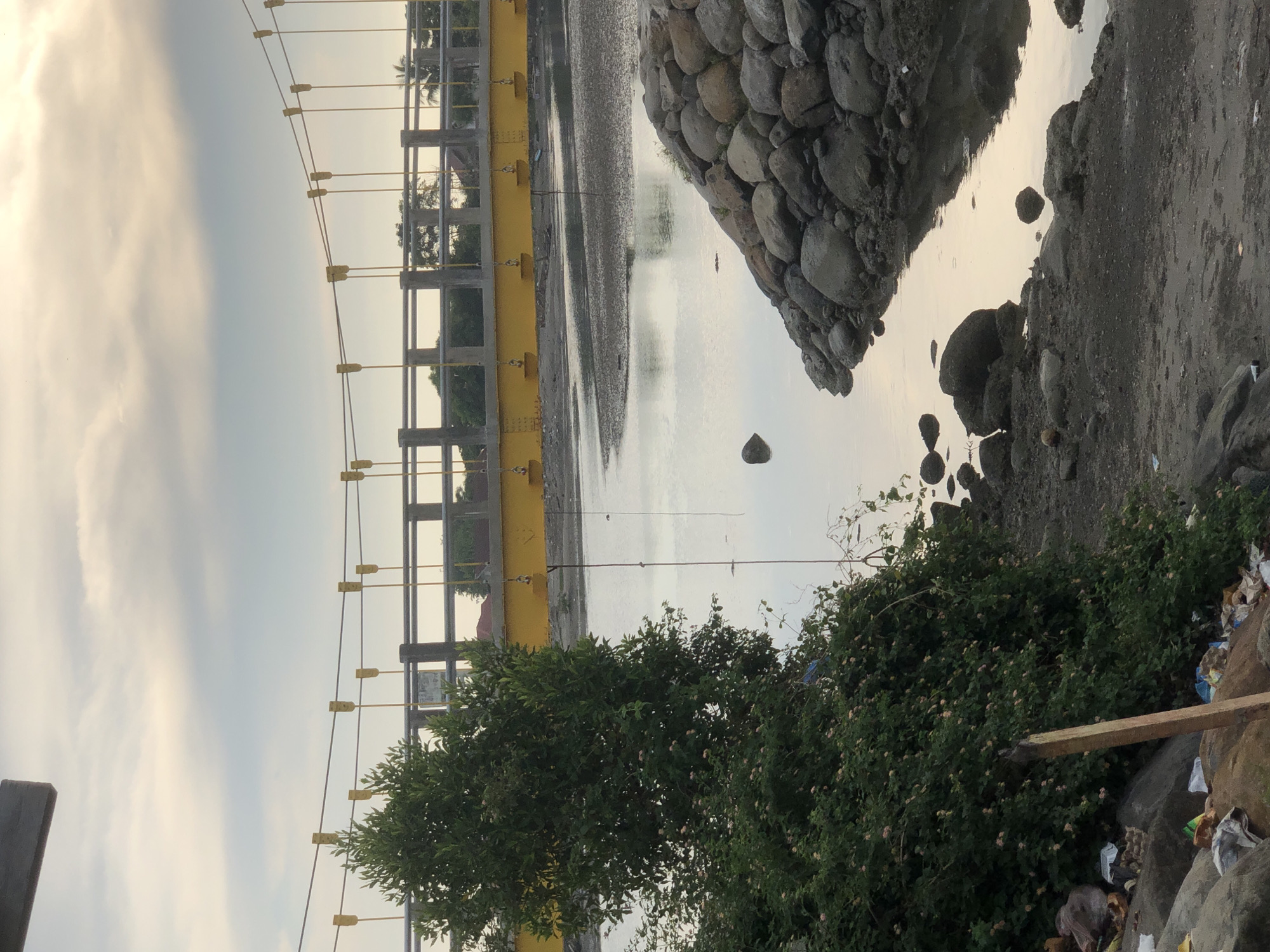 This is a photograph of the Punteut Bridge in Lhokseumawe, Indonesia, taken at dusk. The bridge is a suspension bridge with a bright yellow deck and concrete pillars supporting it. The cables are a light grey, and yellow attachments are visible along the cables. The sky above is overcast with soft, diffused light.
In the foreground, a dense green bush with small pink flowers dominates the left side of the frame. Some trash and debris are visible on the ground near the bush. To the right of the bush, a body of water reflects the muted colors of the sky. The water's edge is lined with a barrier of large, rounded grey stones. Some smaller stones are scattered in the water and on the muddy bank. The background beyond the bridge shows a treeline with palm trees and hints of buildings. The overall scene suggests a somewhat urban or semi-urban coastal environment.