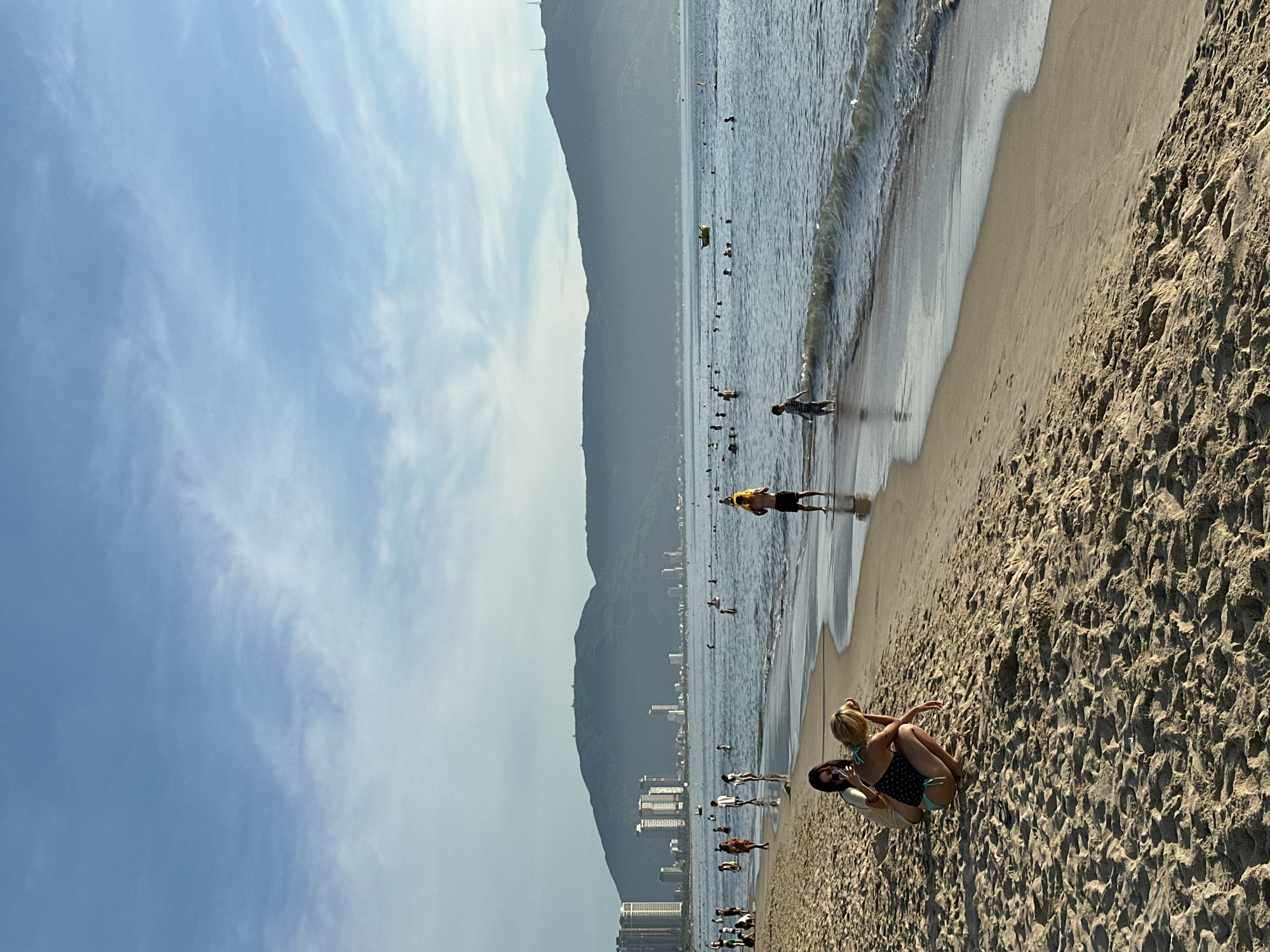 The image depicts My Khe Beach in Da Nang, Vietnam. The foreground is dominated by the sandy beach with footprints and ripples. In the midground, two women are sitting on the sand, one of whom is taking a photo with her phone. Further out, the ocean is populated with numerous people swimming and wading in the gentle waves. A lifeguard in a yellow shirt stands near the water's edge. A young boy is walking towards the shore from the water. In the background, a hazy mountain range rises behind the city skyline, characterized by several high-rise buildings and a few radio towers. A small yellow boat is visible on the water in the distance. The sky is partly cloudy with a light blue hue.