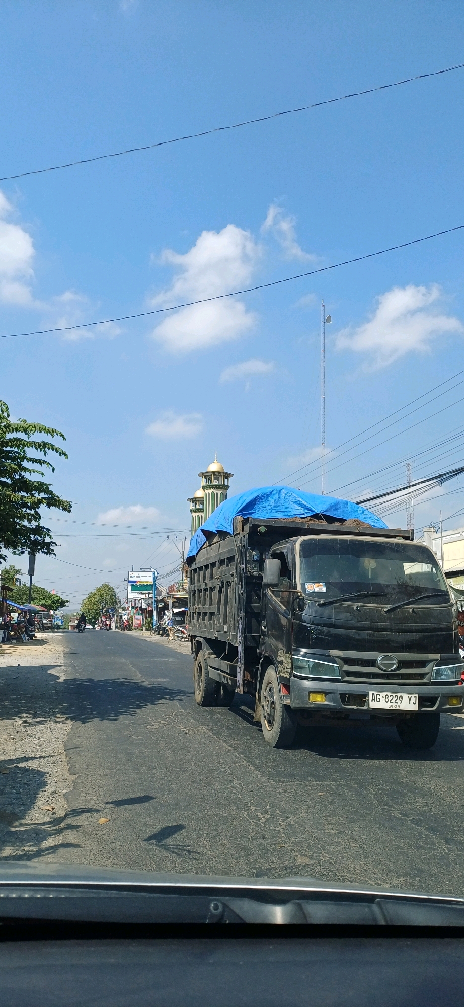 The image captures a frontal view of a black dump truck driving on a paved road under a bright blue sky with scattered clouds. The truck is loaded with dirt and covered with a bright blue tarp. The license plate of the truck reads "AG-8229 YJ." In the background, two mosque minarets with golden domes are visible, characteristic of a mosque. The surrounding environment appears to be a semi-urban or rural area, with a few buildings and trees lining the road. A telecommunications tower stands tall in the distance. The road has some cracks and wear. The foreground shows the dashboard of a vehicle, suggesting the photo was taken from inside a car. This scene is consistent with a street view in Pucakwangi, Indonesia, likely on a clear day.