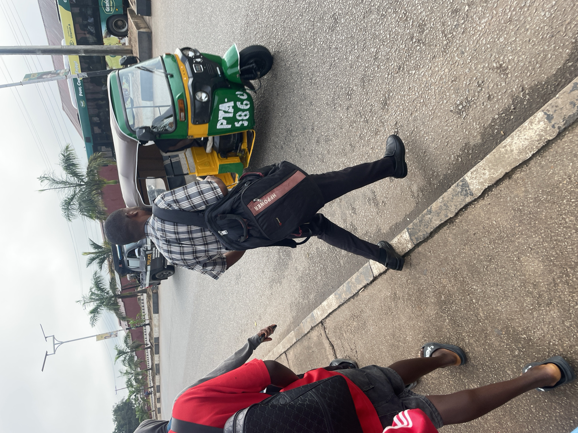 A man in a plaid shirt and dark pants walks on a paved road in Jos, Nigeria, carrying a black messenger bag. To his left, a green and yellow auto-rickshaw is parked. In the background, a green bus with "Grand" written on it is visible, as is a police truck. Palm trees and a street light line the side of the road. Another person in a red shirt and dark shorts is visible in the foreground, walking away from the camera.
