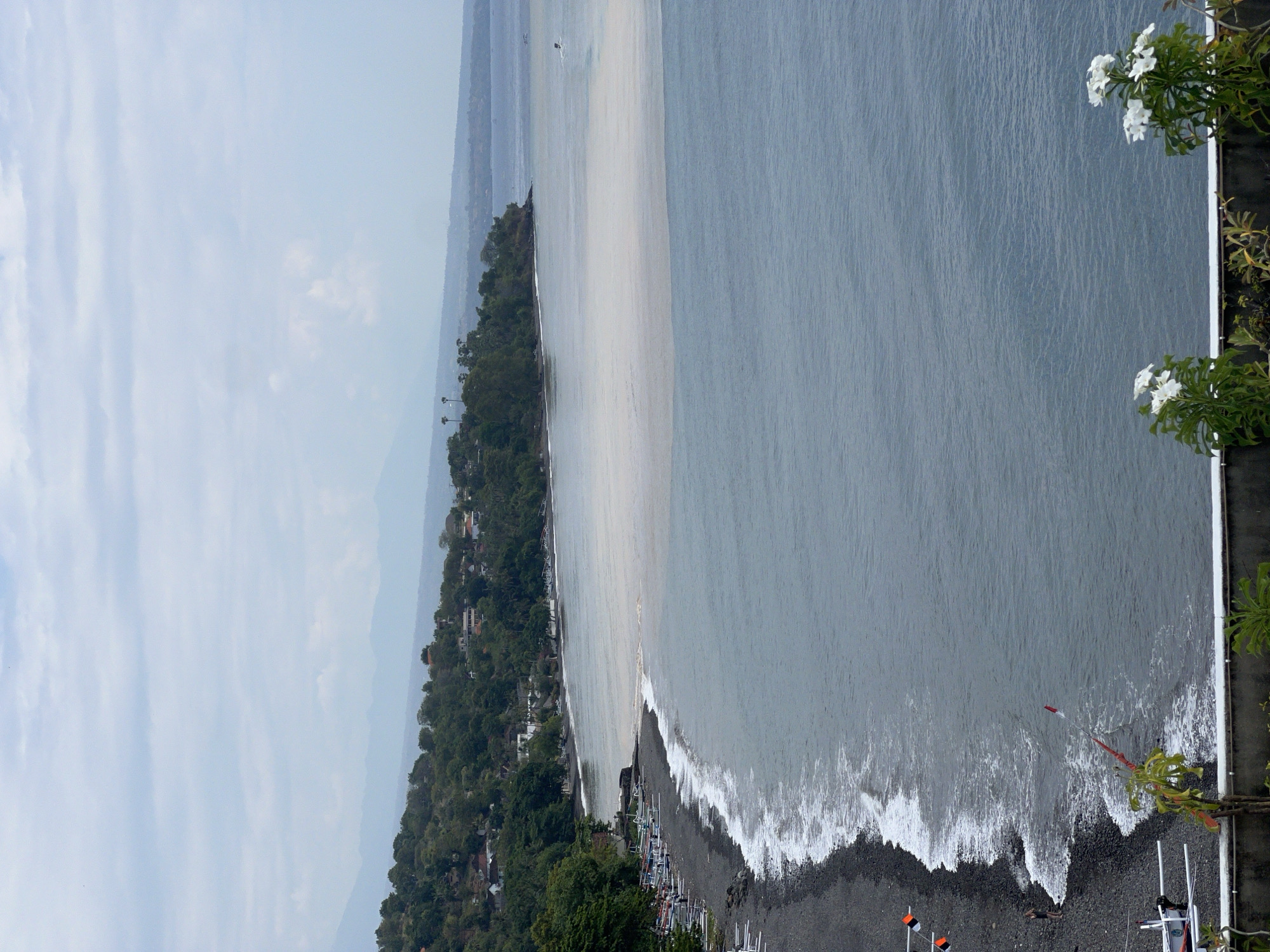 This is an aerial view of a coastal scene in Abang, Indonesia. The image depicts a dark, pebbly beach with waves gently rolling onto the shore. Numerous outrigger boats are lined up along the beach. The water transitions from a lighter, cloudy hue near the shore to a deeper blue further out. A green, tree-covered hillside with scattered buildings forms the backdrop along the coastline. The sky is overcast with soft clouds. In the foreground, a white wall and some white flowers are visible. A person is standing on the beach near the water's edge. There are also flagpoles with flags and what appear to be parts of boats in the very front of the image. The overall scene conveys a sense of a coastal village.