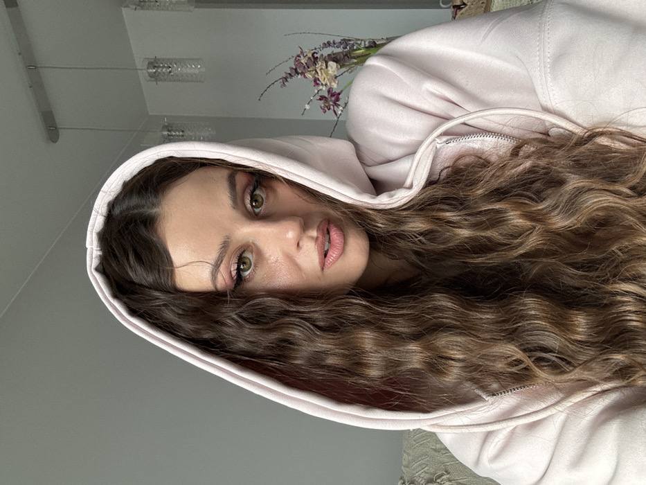 The image displays a close-up, waist-up portrait of a female individual positioned indoors in a residential setting in Krasnoyarsk, Russia. The subject has long, wavy dark brown hair and light-colored eyes, with visible pink eyeshadow and eyeliner. She wears a light pink hooded sweatshirt; the hood is up and frames her face, and a silver zipper is partially visible on the garment's front. Her gaze is directed towards the camera with a neutral expression. The background features light-toned walls, a white ceiling, and three suspended modern pendant light fixtures with clear, textured glass shades on the right side. A section of light-colored, textured upholstery or fabric is visible on the far left, and dried purple and brown floral arrangements are present in the upper right background.