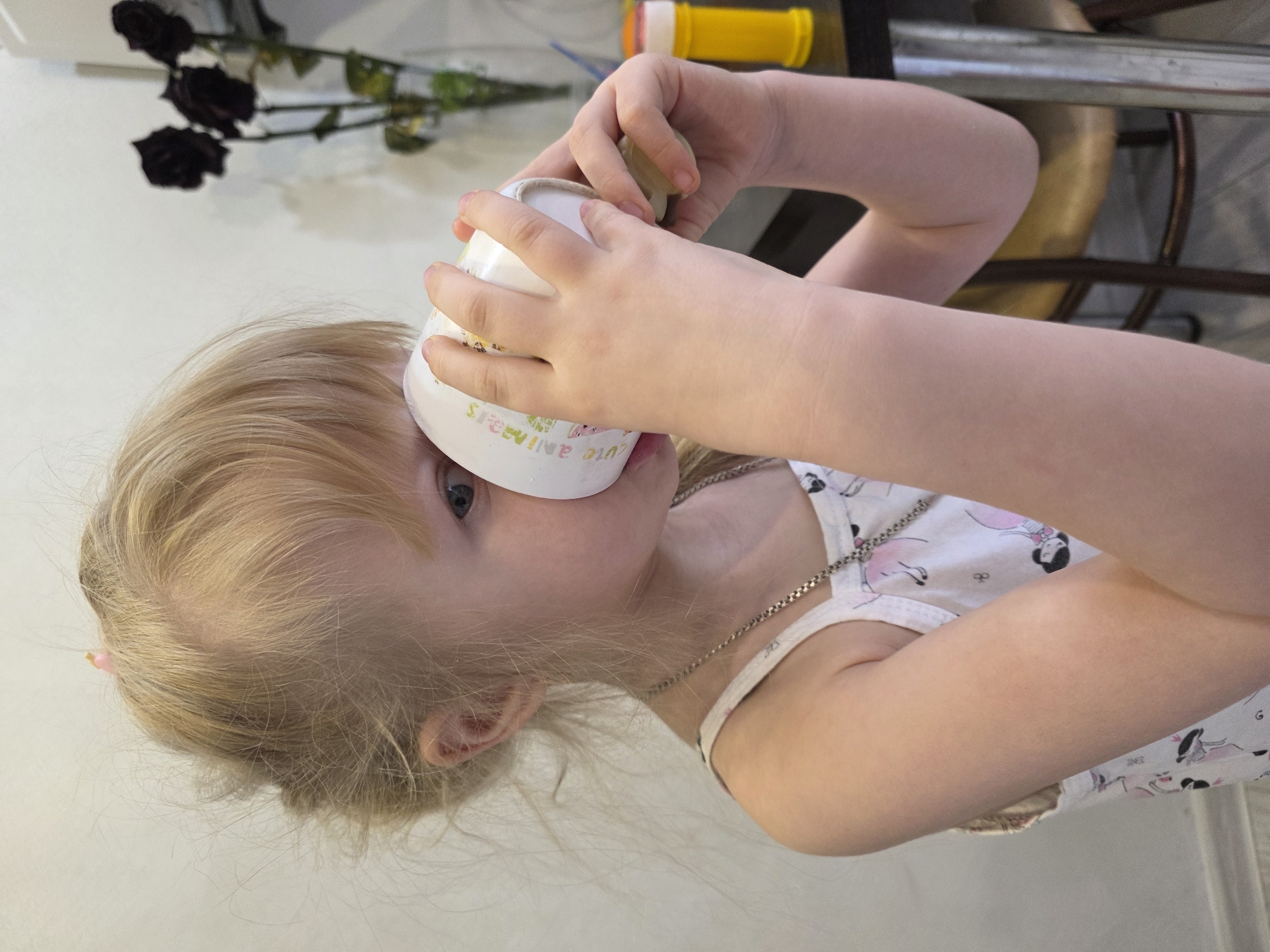 A child with blonde hair is drinking from a white cup with colorful letters, wearing a white tank top with princess drawings and a silver necklace. The child is in a room that includes decorative black flowers, a yellow and orange object, and a metal chair. The location is Chaikovsky, Russia.