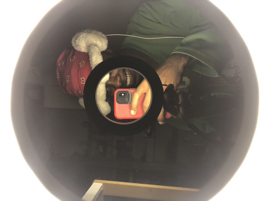 An individual is depicted in a mirror selfie, taken indoors in Tema, Ghana. The subject, a person with dark skin, is smiling broadly, revealing teeth. They are wearing a red bandana-style cap with white patterns, featuring white faux-fur ear flaps. A green long-sleeved shirt is also visible. The individual is holding a red-cased smartphone (likely an iPhone) which is capturing the image. A black circular ring light is positioned directly in front of the smartphone, its reflection dominating the center of the frame and illuminating the subject's upper face. The subject's left hand is visible through the ring light, holding the smartphone. A thin white cable extends from the right side, possibly connected to the ring light or an earphone. The background, reflected in the mirror, is dimly lit and shows indistinct shapes, including what appears to be light-colored shelving or furniture on the left. The ring light provides the primary illumination for the scene.