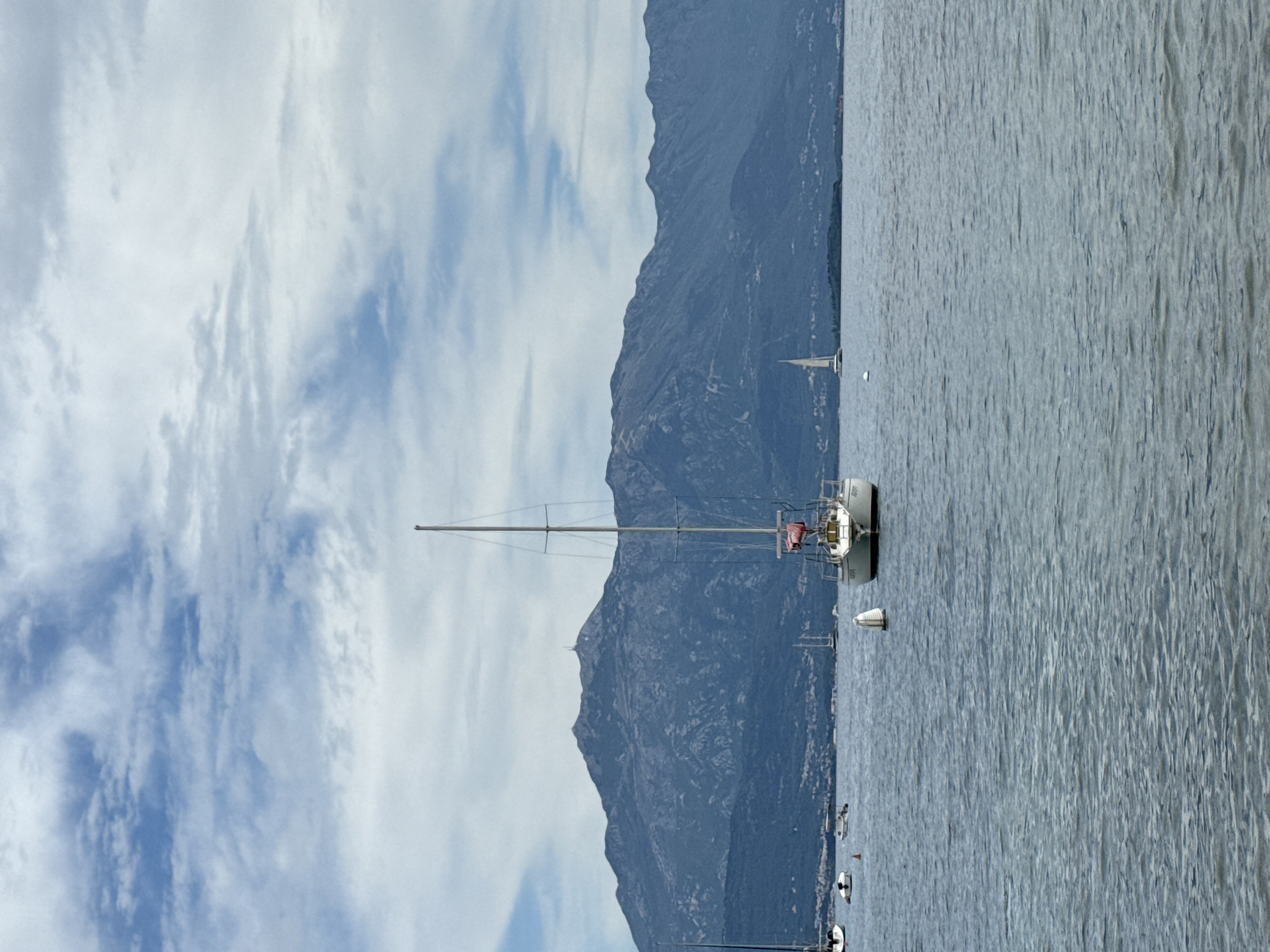 The image shows a sailboat anchored in the water, with a mountain range in the background. The sky is cloudy with patches of blue. The water is calm with slight ripples. Đenovići, Montenegro, cannot be determined from the image provided.
