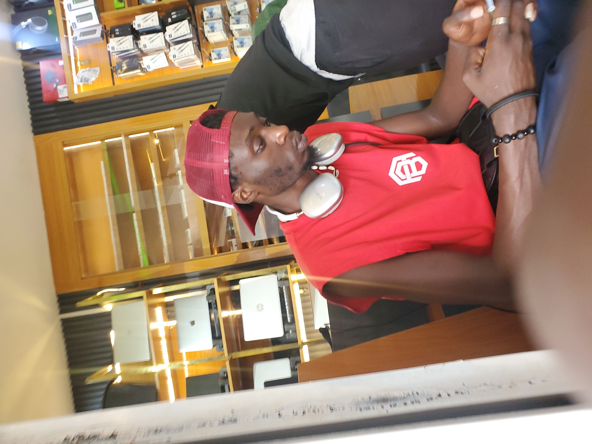 The image shows a young Black man wearing a red sleeveless t-shirt and a red baseball cap. He has headphones around his neck and is looking down, holding a cigarette in his right hand. He is wearing a bracelet on his left wrist and rings on two fingers of his right hand. The background appears to be a store with shelves displaying electronic devices, including laptops. The scene is well-lit, suggesting an indoor environment. The man is seated, and part of another person in a dark shirt is visible behind him. The image composition suggests it was taken from a low angle, possibly from a seated position by the photographer. The background shelves and displays strongly suggest the location is a retail space for electronics, potentially in a commercial district of Jos, Nigeria.