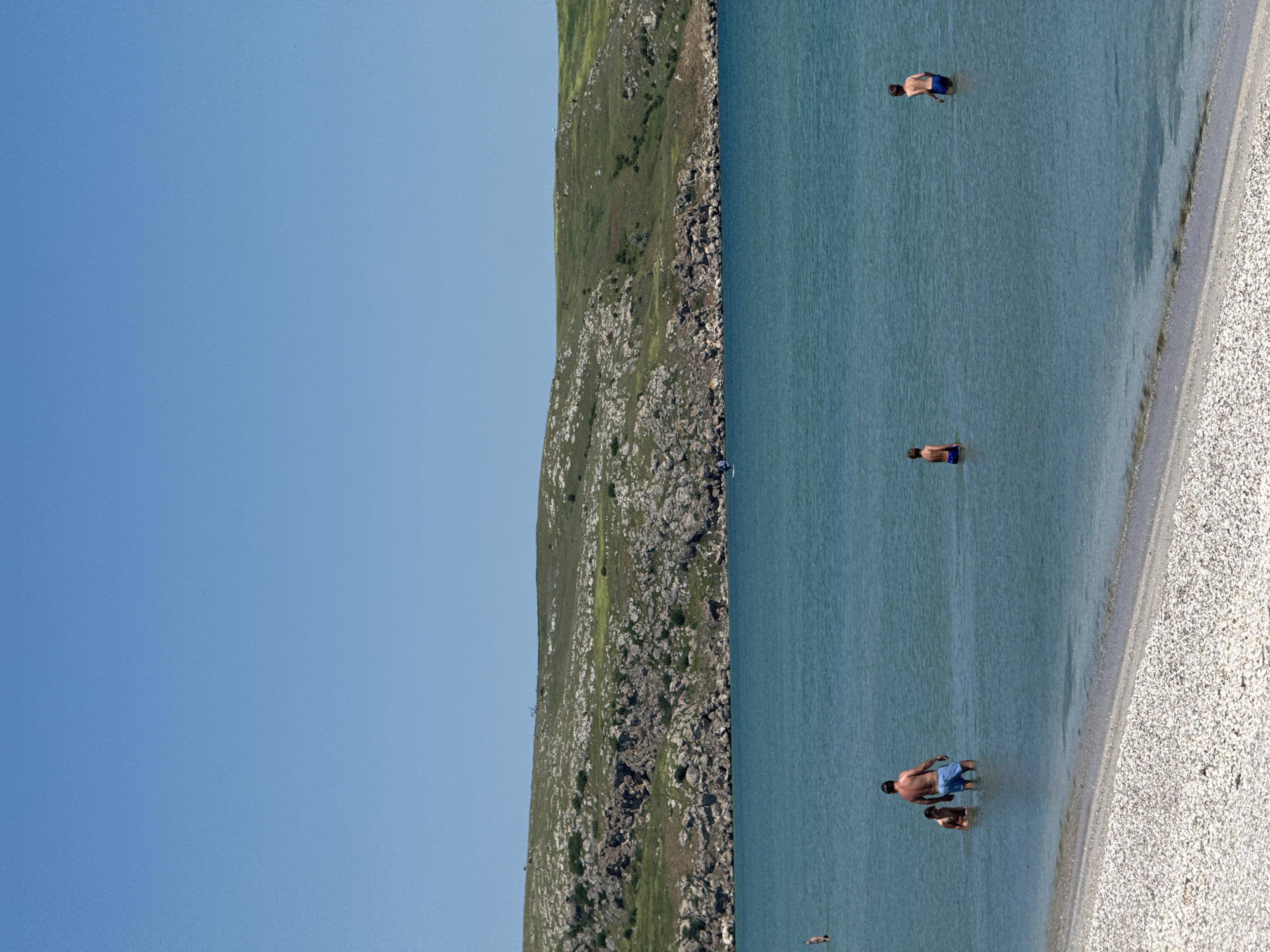 The image shows a beach scene in Мысовское сельское поселение, Ukraine. The camera is positioned facing the sea with a pebble beach to the lower right of the frame. The water is a clear turquoise color. Three individuals are seen swimming in the water at varying depths. Along the left edge of the image, there is a rocky, grassy hill that runs vertically. The sky is a clear, light blue. The image is rotated 90 degrees counterclockwise.