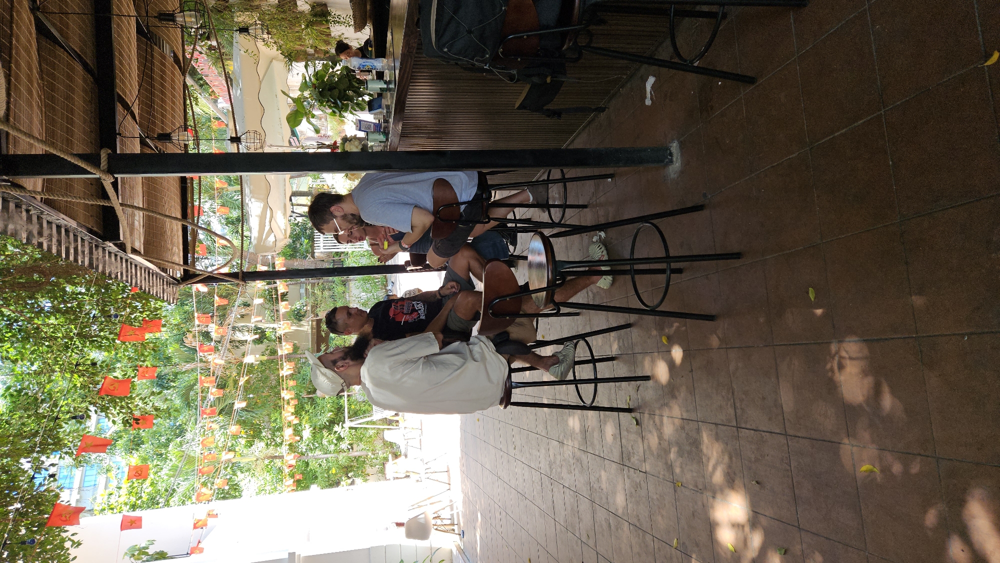 **Scene:** Outdoor patio of a bar or cafe in Nha Trang, Vietnam.
**People:** Three men are seated on tall stools along a bar counter. One man wears a light blue shirt, another a black t-shirt, and the third a white shirt. They are engaged in conversation.
**Objects:** Bar stools, bar counter, red communist flags strung overhead, lush greenery, brown tiled floor.
**Environment:** The area is shaded and the scene is illuminated by natural light. The location seems to be a tourist area.