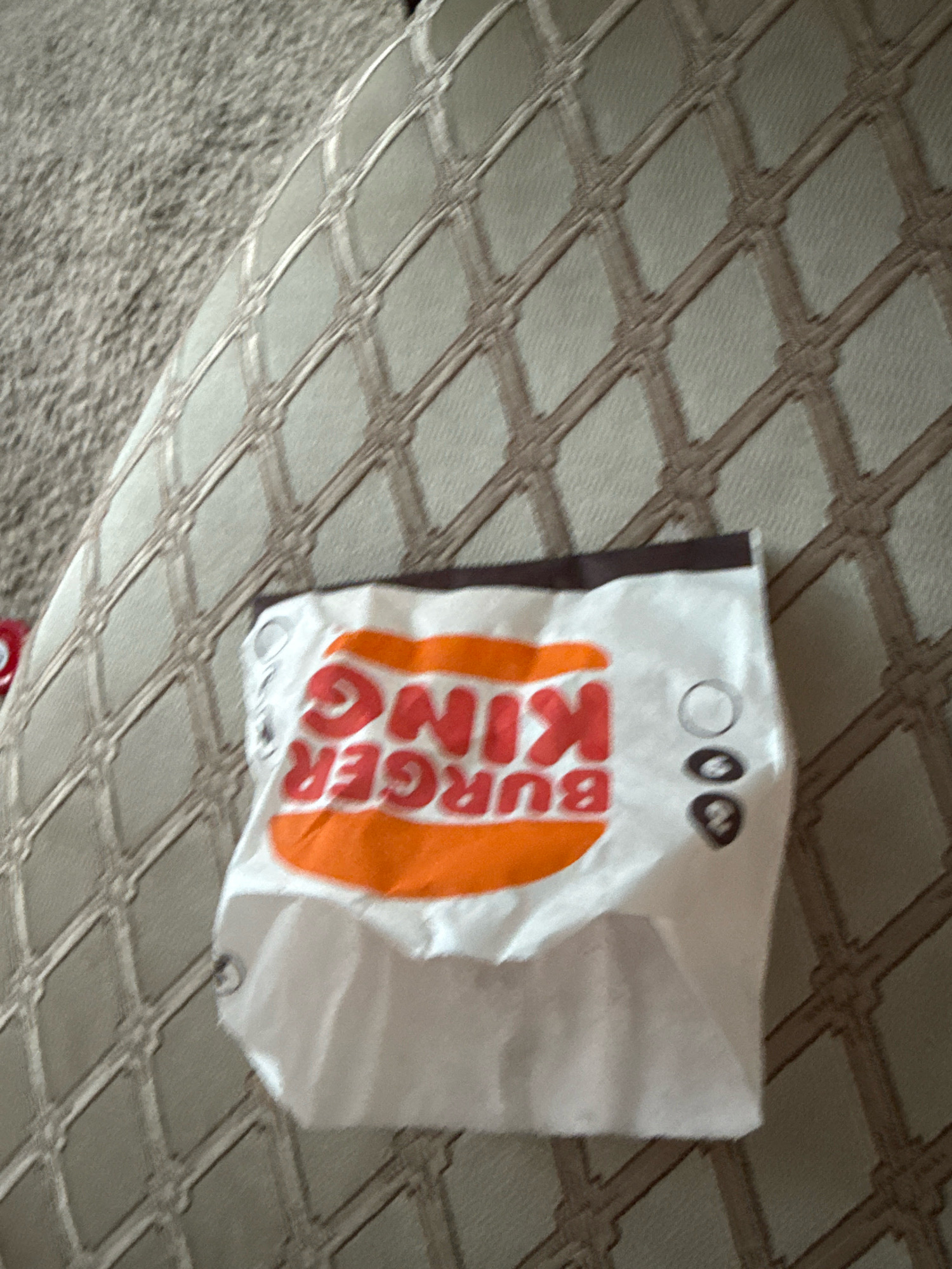An empty Burger King bag is lying crumpled on a textured beige surface, likely a couch or upholstered chair. The bag's white exterior features the prominent red Burger King logo with orange accents, reading "BURGER KING" upside down. To the right of the logo, there are three small, dark circular markings. The background is a grey, slightly blurred carpet. There are no people present in the image. The scene is indoors and appears to be a casual setting.