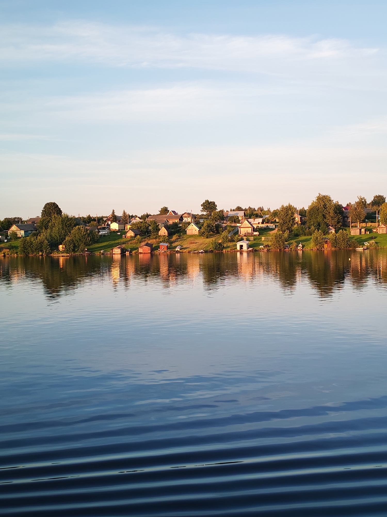 The image depicts a serene lakeside scene in the village of Bol'shaya Stepanovskaya, Russia. The foreground is dominated by the calm, reflective surface of a lake, with subtle ripples distorting the mirrored image of the opposite bank. The far bank of the lake is lined with a collection of small, rustic houses and sheds, nestled amongst lush green trees and vegetation. The houses appear to be primarily single-story structures, with pitched roofs, some painted in light colors and others made of wood. Several small boats are visible near the shore, suggesting a fishing or leisure community. The sky above is a soft gradient of pale blue and pink, indicative of either early morning or late evening light, with wispy clouds scattered across it. The overall impression is one of peaceful, rural tranquility.