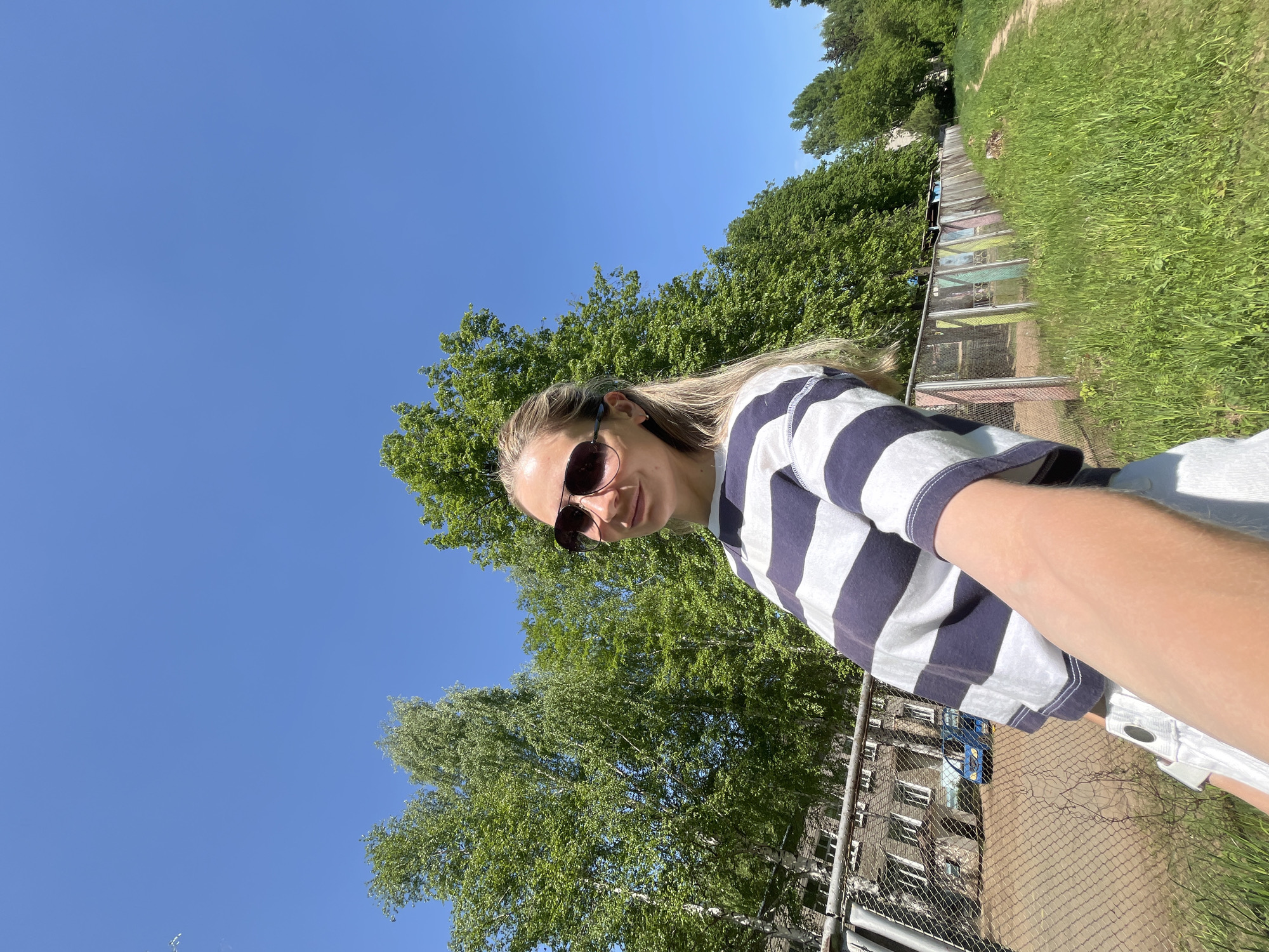 A medium shot shows a woman wearing sunglasses and a blue-and-white striped shirt, posing for a selfie outdoors in the city of Chaikovsky, Russia. The woman is positioned slightly off-center to the left, with a bright blue sky visible above and to the left of her. Behind her is lush greenery consisting of trees and grass. A long, partially obscured structure resembling a greenhouse or a series of cages is also visible. In the lower part of the frame, a chain-link fence and a multi-story brick building can be seen. The lighting suggests a sunny day. The camera angle is slightly tilted, giving an angled perspective of the scene.