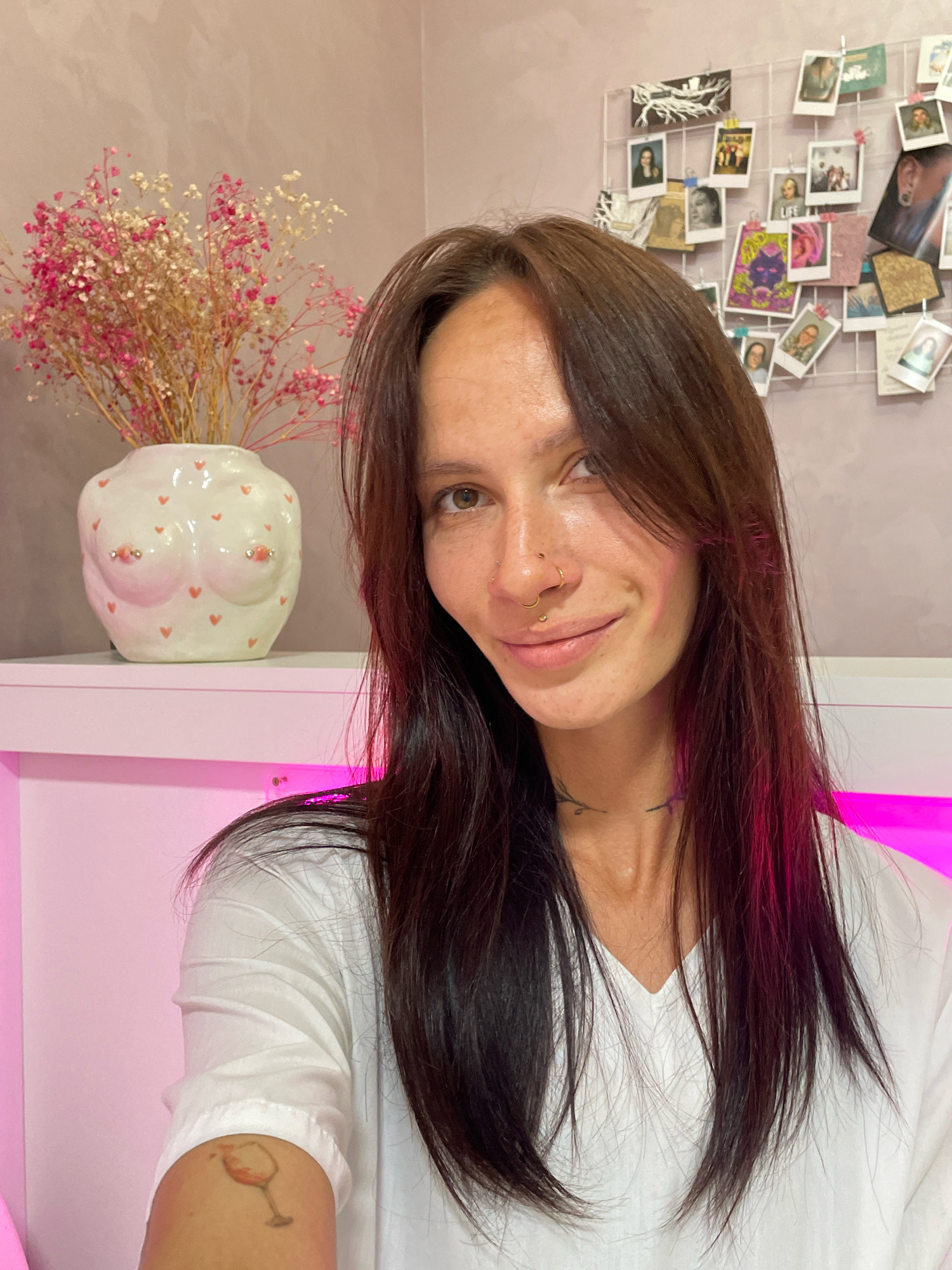 This is a medium-shot, portrait-style photograph of a young adult female with long, dark brown hair, facing the camera and smiling. She is wearing a white v-neck t-shirt. She has a nose ring and a septum piercing. A tattoo of a wine glass is visible on her upper left arm. In the background, to the left, is a white ceramic vase shaped like breasts, adorned with small pink hearts, holding dried pink and white flowers. To the right, a wire grid is covered with various small photographs and cards. The overall setting appears to be an indoor environment with a pink-toned wall and a bright pink light source illuminating the lower right portion of the image. There is no information available to determine the location in Izhevsk, Russia.
