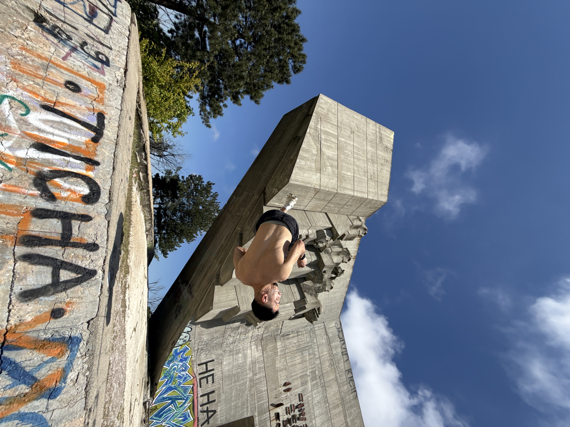 A man is hanging upside down on a concrete structure in Varna, Bulgaria. He is shirtless, wearing black shorts and white socks. The structure is weathered and has graffiti on its surface. The sky is blue with scattered clouds. Trees are visible in the background. The photo is taken from a low angle.