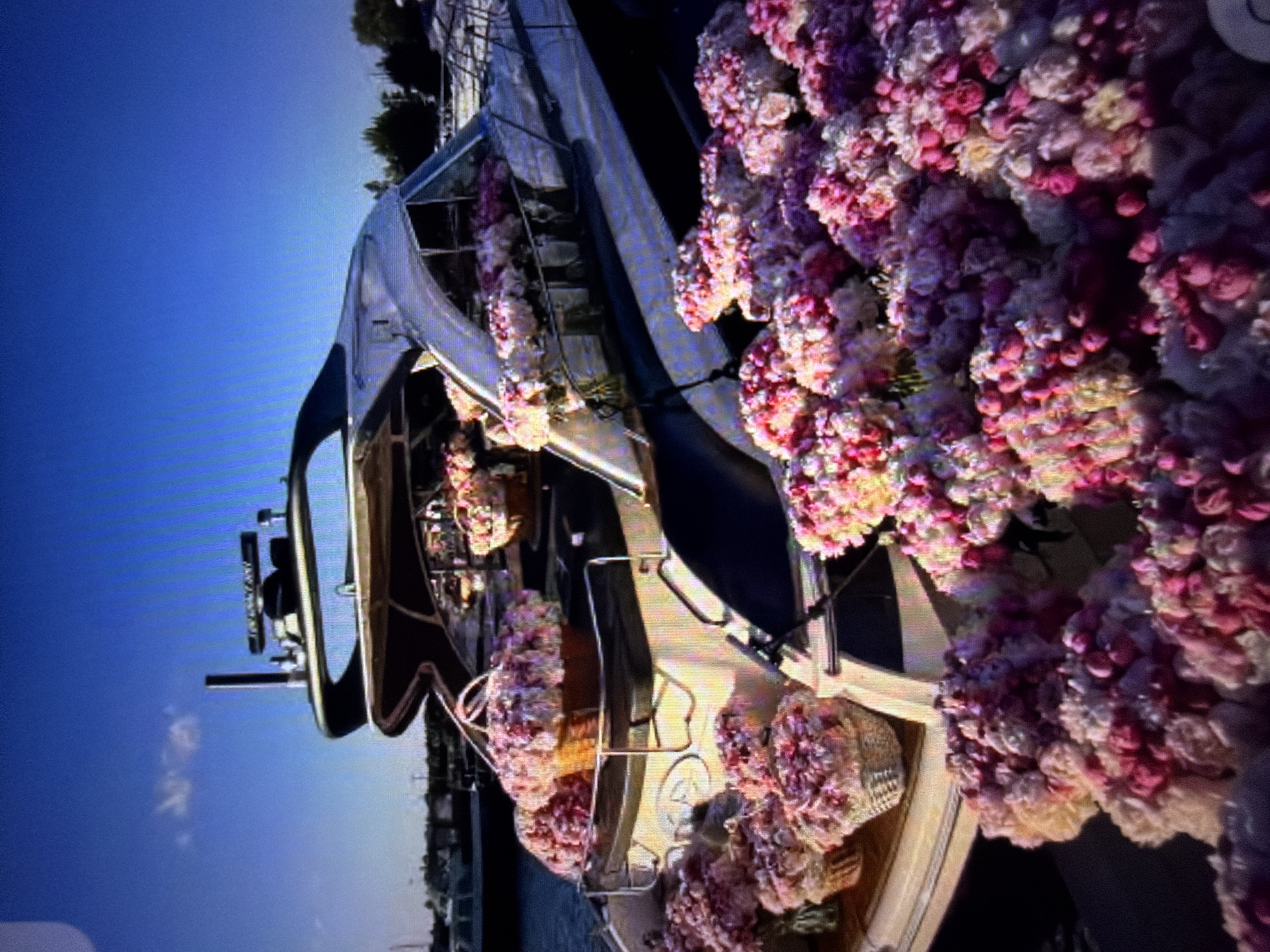 A yacht, heavily adorned with pink and purple floral arrangements, is docked in a body of water. The flowers are clustered together, forming dense bouquets and cascades that drape over the boat's railings and deck. Some of the floral arrangements are in woven baskets. The yacht appears to be a luxury vessel with multiple levels and a sleek, white exterior. In the background, a clear blue sky with a few wispy clouds suggests fair weather and daytime. The setting appears to be a marina or harbor, with other boats and structures visible in the distance, possibly indicating a coastal or riverside location. The overall impression is one of an elaborate celebration or event, possibly a wedding or party, taking place on the water. The vibrant colors of the flowers create a festive and romantic atmosphere. There is no visible text in the image.