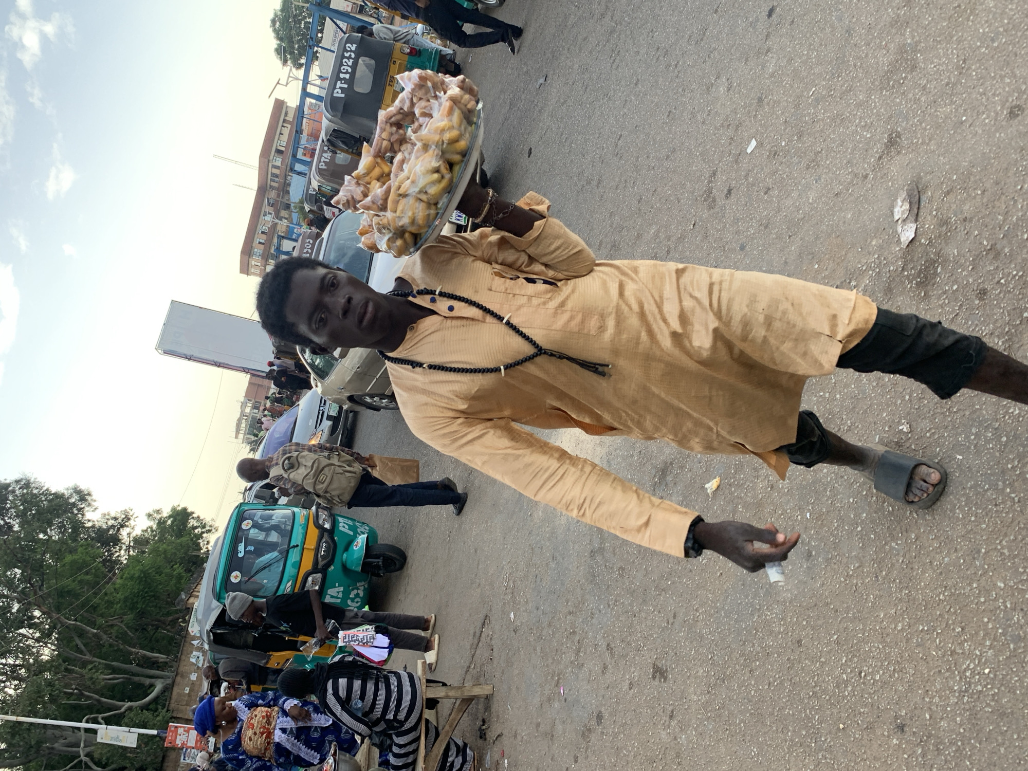 A young Black male vendor in Jos, Nigeria, walks towards the camera, carrying a large tray of fried snacks on his right arm. He wears a long-sleeved, tan-colored traditional tunic and black pants, with black sandals on his feet. A beaded necklace hangs around his neck. In his left hand, he holds what appears to be a folded wad of currency. The vendor is on a paved street with scattered debris.
Behind him, a teal and yellow auto-rickshaw is parked, with a male driver visible. Further back, other vehicles, including cars and auto-rickshaws, are present, suggesting a busy urban environment. A group of people, including women in brightly colored clothing and a person wearing a backpack, are visible on the left side of the frame, near a wooden bench. Trees and buildings form the background under a hazy sky. The scene appears to be a street market or a busy thoroughfare in Jos.