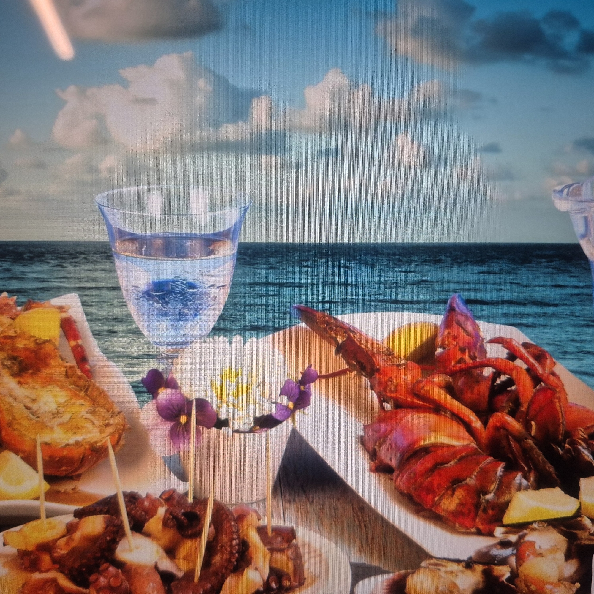 The image depicts a table set with a seafood meal and a glass of water against a backdrop of the ocean and sky. On the table, there is a cooked lobster, a dish of what appears to be octopus, and a glass filled with clear liquid. The meal is garnished with lemon wedges and small purple flowers. The ocean stretches out in the background, meeting a sky filled with fluffy white clouds. The scene is set outdoors, likely near the coast. There are no people visible in the image. There is no information available about the location in the city of Nowa Wieś Książęca, Poland.