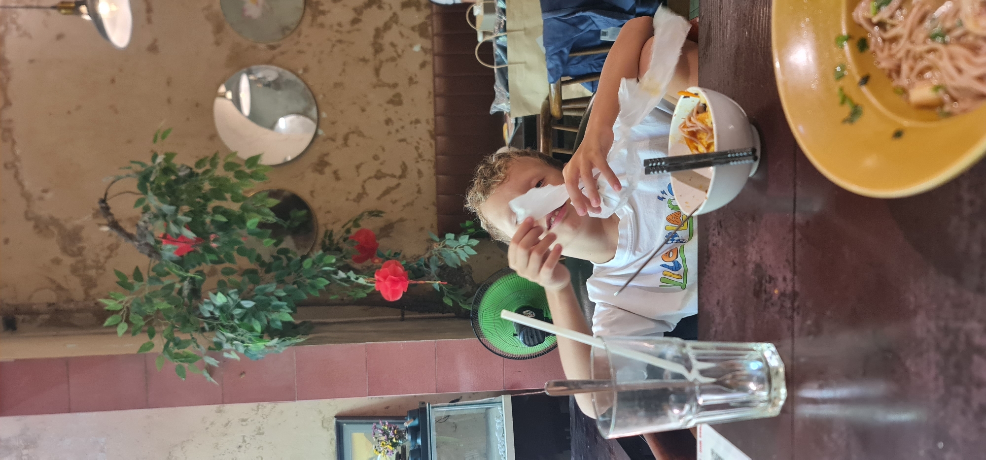 A boy is at a table in a restaurant in Đà Nẵng, Vietnam. He is seated, wearing a white shirt with colorful designs. He holds a tissue to his face with his left hand. In front of him are two bowls. One is yellow and contains noodles. The other is white, and has chopsticks resting on top. To his left, there is a green fan and a glass of water with a straw. The restaurant’s decor includes mirrors on the wall and a plant with red flowers.