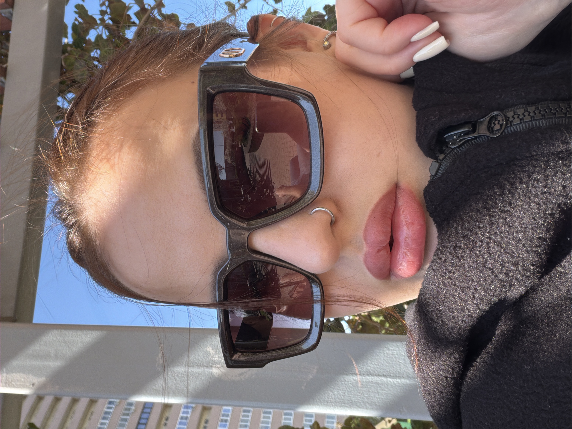 A close-up, rotated image shows a person wearing sunglasses and a nose ring. The sunglasses are dark and have a thick frame with a detail at the top corner. The person is wearing a dark-colored, zipped-up jacket. The background reveals a building with multiple windows and blue sky, suggesting an outdoor setting in Krasnodar, Russia.