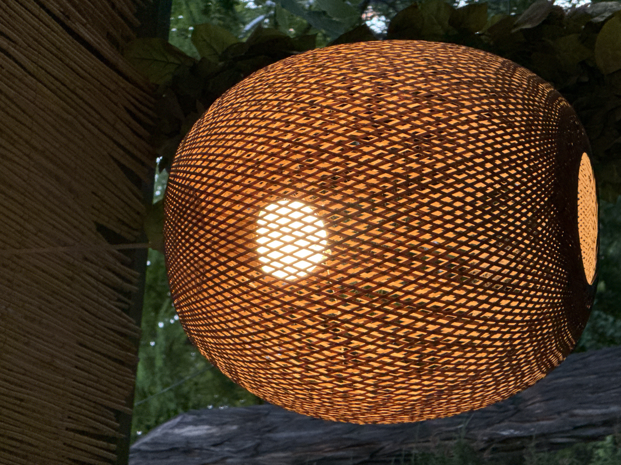 A spherical woven light fixture, with an internal light source illuminated, is suspended outdoors. The fixture is positioned in front of foliage and a structure made of stacked, thin wooden rods, possibly part of a pavilion or gazebo. There is a glimpse of a dark, possibly wooden or stone, structure below the lamp. The photo's location is in Tashkent, Uzbekistan.