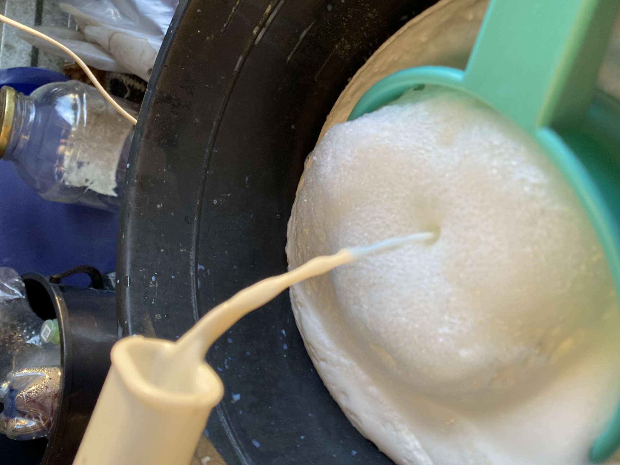In Maloarslangulovo, Russia, a close-up shot captures a pale yellow liquid being poured from a beige plastic spout into a black plastic basin filled with a frothy white substance. The spout is angled downwards, with the liquid forming a stream that disappears into the foam. The white substance appears to be a mixture of liquid and air, creating a dense, bubbly layer that fills a significant portion of the basin. A teal-colored handle, possibly part of a sieve or strainer, is partially submerged in the foam at the lower right of the frame. In the background, a glass jar with a metallic lid is visible, along with other objects that are out of focus, suggesting an outdoor or semi-outdoor setting. The overall scene suggests a process of mixing or preparation involving liquids and foam.