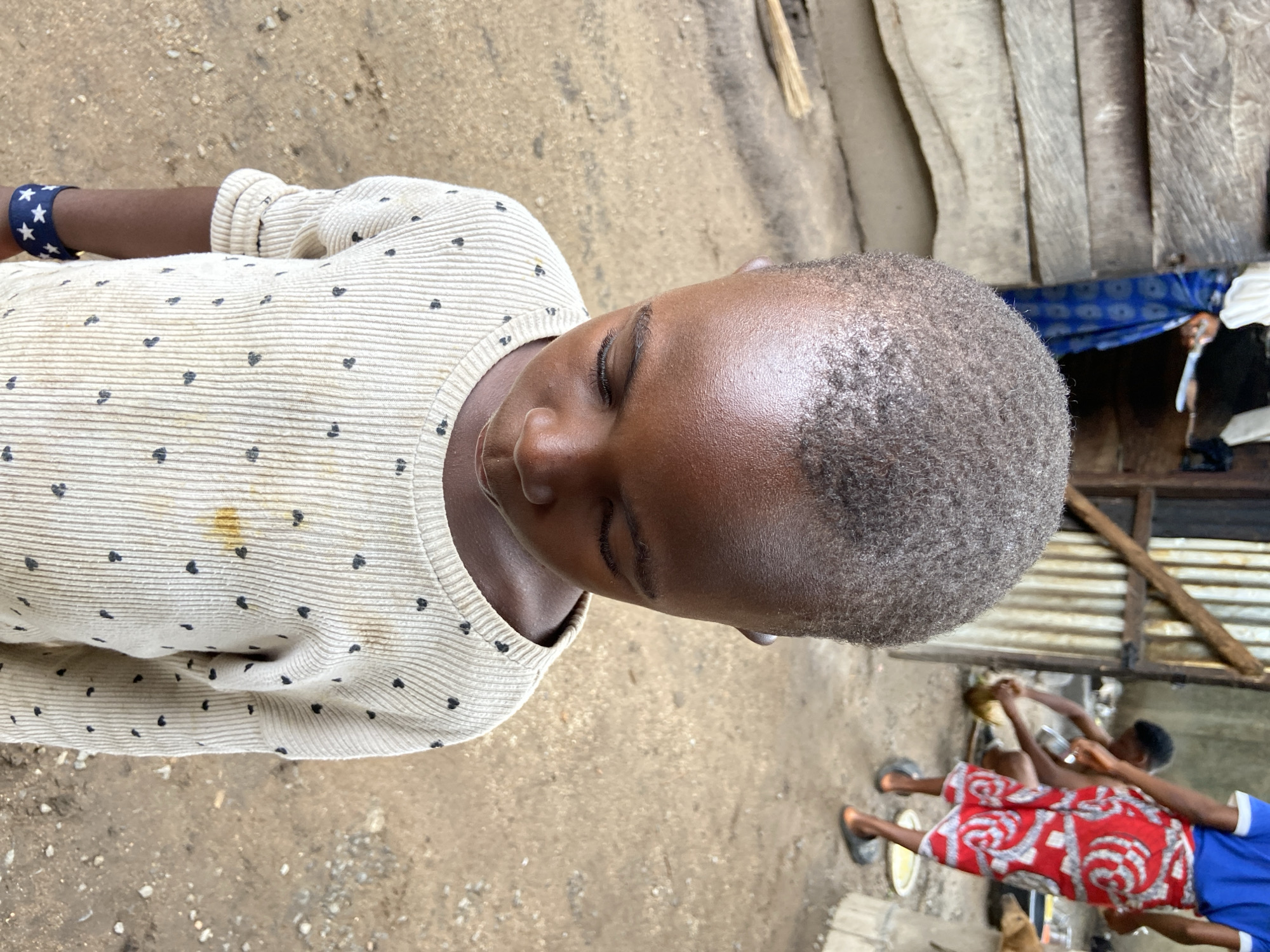 A photo depicts a child with dark skin and short hair wearing a cream-colored sweater with small, dark heart patterns. They are positioned against a dirt ground, and their eyes are closed. A dark blue bracelet adorned with white stars is visible on their left wrist. The sweater has a stain on the front.
In the background, two people wearing red and blue outfits are discernible near a structure made of wooden planks. It is not possible to accurately discern the location in the city of Mararaba, Nigeria. The background appears to be an outdoor environment.