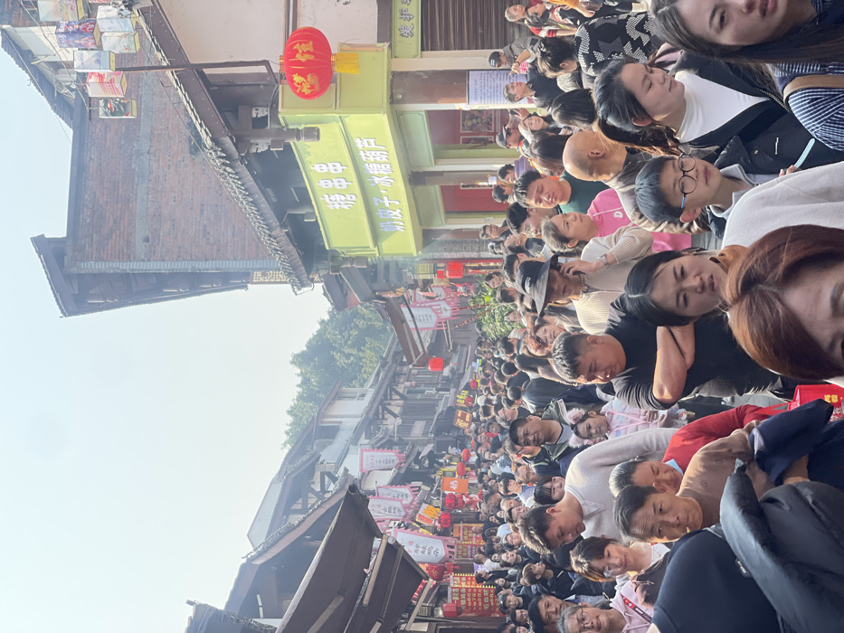 A crowded street scene unfolds in what appears to be a traditional Asian town. Buildings with pitched roofs and decorative lanterns line the narrow street. A sea of faces, mostly Asian, fills the space between the buildings. People are packed tightly together, with some looking towards the camera and others focused on something further down the street. A few red lanterns hang overhead, adding pops of color. The overall impression is of a bustling market or festival atmosphere.