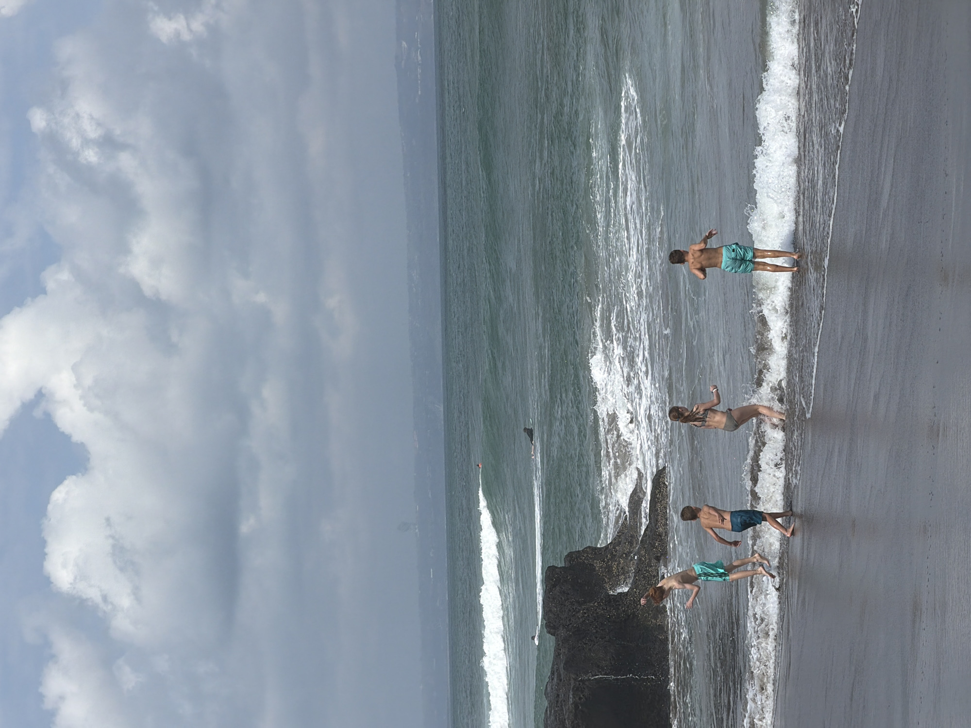 In North Kuta, Indonesia, on a beach with dark sand, four children are playing near the water's edge. Two boys and two girls are present. One boy, with red hair, is in mid-air, jumping with his arms outstretched. He wears turquoise swim trunks. Beside him, another boy in dark blue swim trunks walks towards the water, his body turned away from the camera. A girl in a bikini stands in the shallow water, splashing. Further to the right, another boy stands in the water, facing away from the viewer, also wearing turquoise swim trunks. The ocean has moderate waves with white foam crashing on the shore. A dark rock formation is visible on the left side of the image, extending into the water. In the distance, several surfers are scattered across the sea, some on boards. The sky is overcast with large, fluffy clouds.