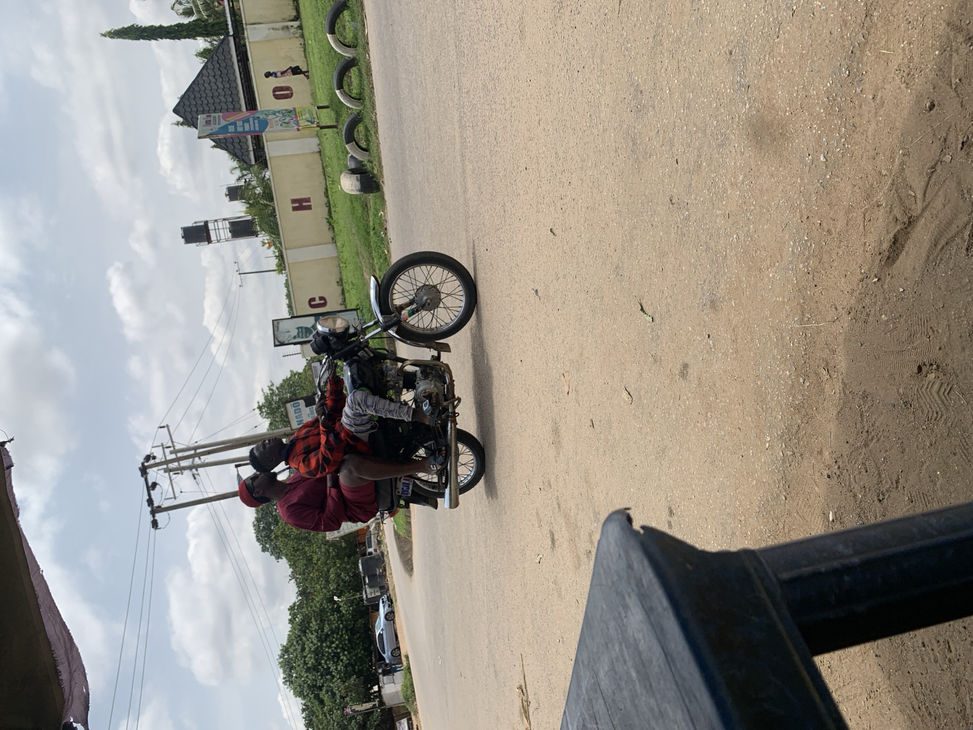 Two individuals, an adult male and a male child, are riding on a motorcycle on a paved road. The adult male, wearing a red outfit and a red cap, is driving the motorcycle. The child, wearing a red and black checkered shirt, is seated behind him. They are in motion, with the motorcycle's wheels visible on the road. The environment appears to be an urban or suburban area with buildings, trees, and power lines in the background. The sky is overcast. There is a dark, weathered table in the foreground, partially obscuring the bottom left of the image. The location is identified as Abuja, Nigeria.