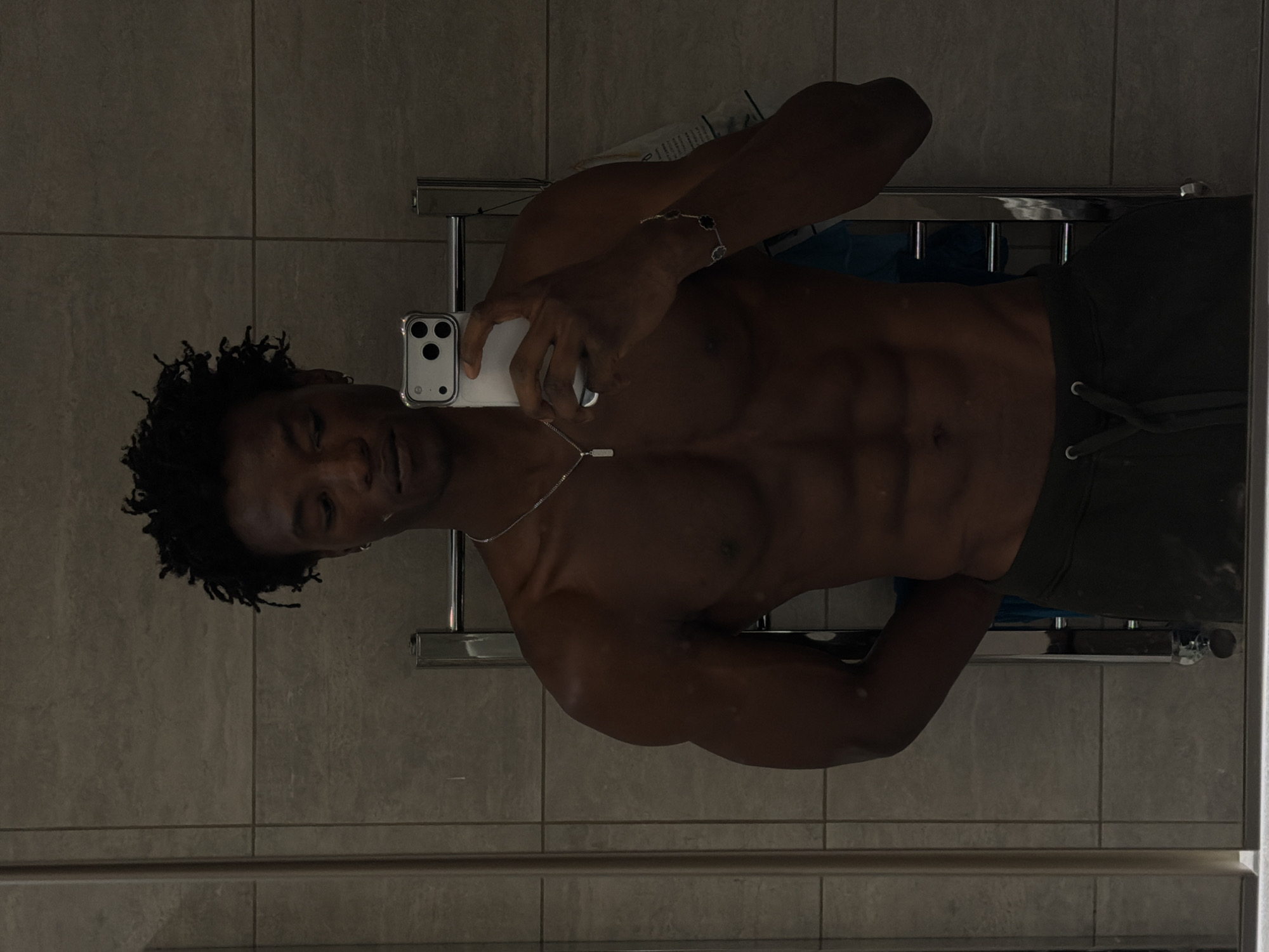 A shirtless Black man with dreadlocks is taking a mirror selfie in a bathroom. He is wearing dark green sweatpants and jewelry, including a silver chain and a bracelet. The bathroom walls are tiled with light grey tiles, and a chrome towel rack is visible behind him. The lighting is dim, casting shadows on his muscular physique.