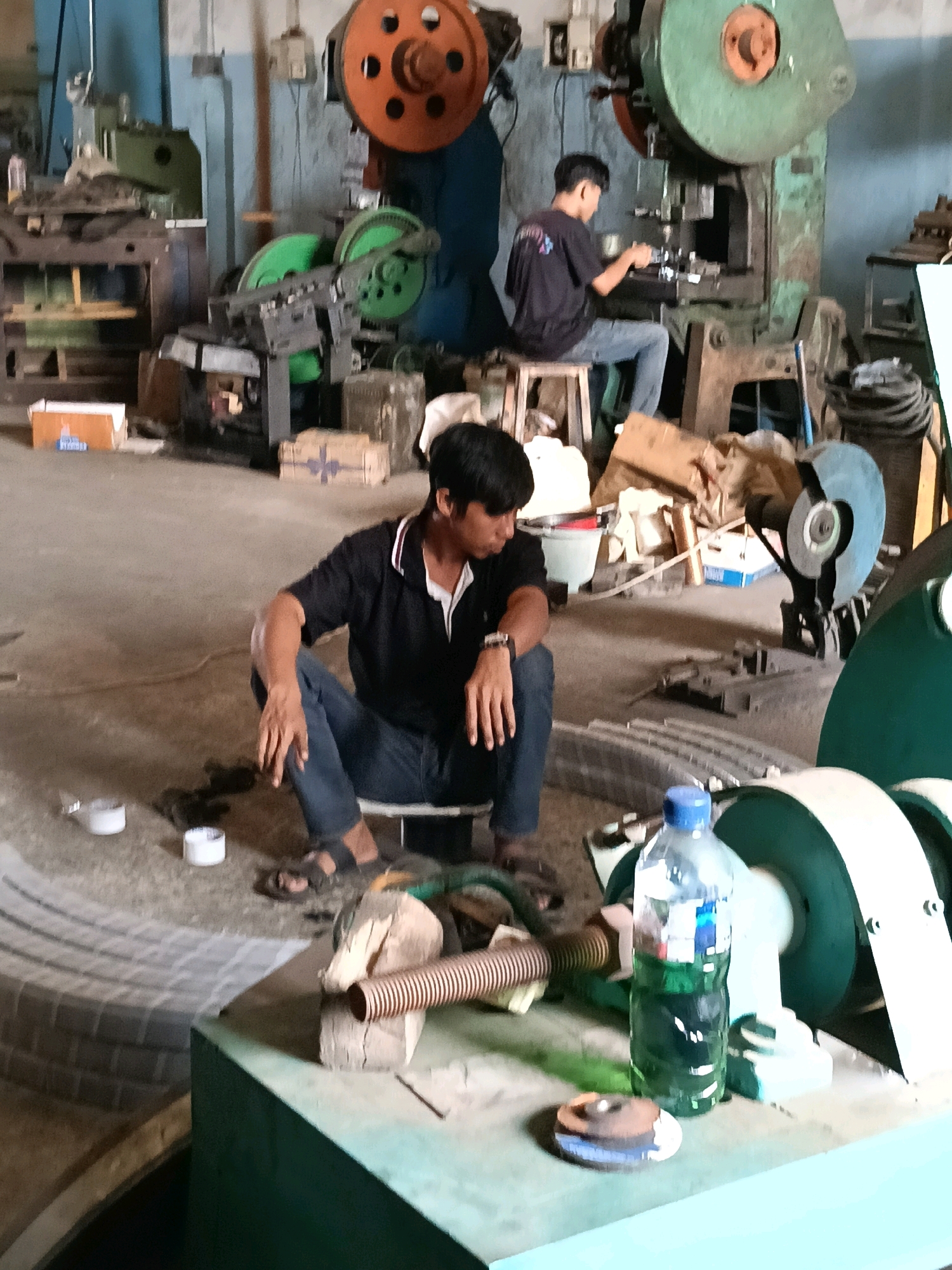 The image depicts a workshop in Belimbing, Indonesia, likely an industrial or repair facility. In the foreground, a young man is seated on the floor, wearing a black polo shirt and blue jeans. He appears to be resting or observing something. Nearby, there are two white cylindrical objects. In the background, another young man is operating a large industrial machine, possibly a press, while seated on a stool. The workshop is filled with various machinery, including large metal presses with distinctive orange and green flywheels, and other industrial equipment. Tools, materials, and boxes are scattered around the space, suggesting an active work environment. The floor is concrete, and the walls appear to be made of a rough material. A bottle of green liquid, likely a coolant or lubricant, is visible on a workbench in the foreground. The overall impression is of a functional, albeit somewhat cluttered, industrial setting.