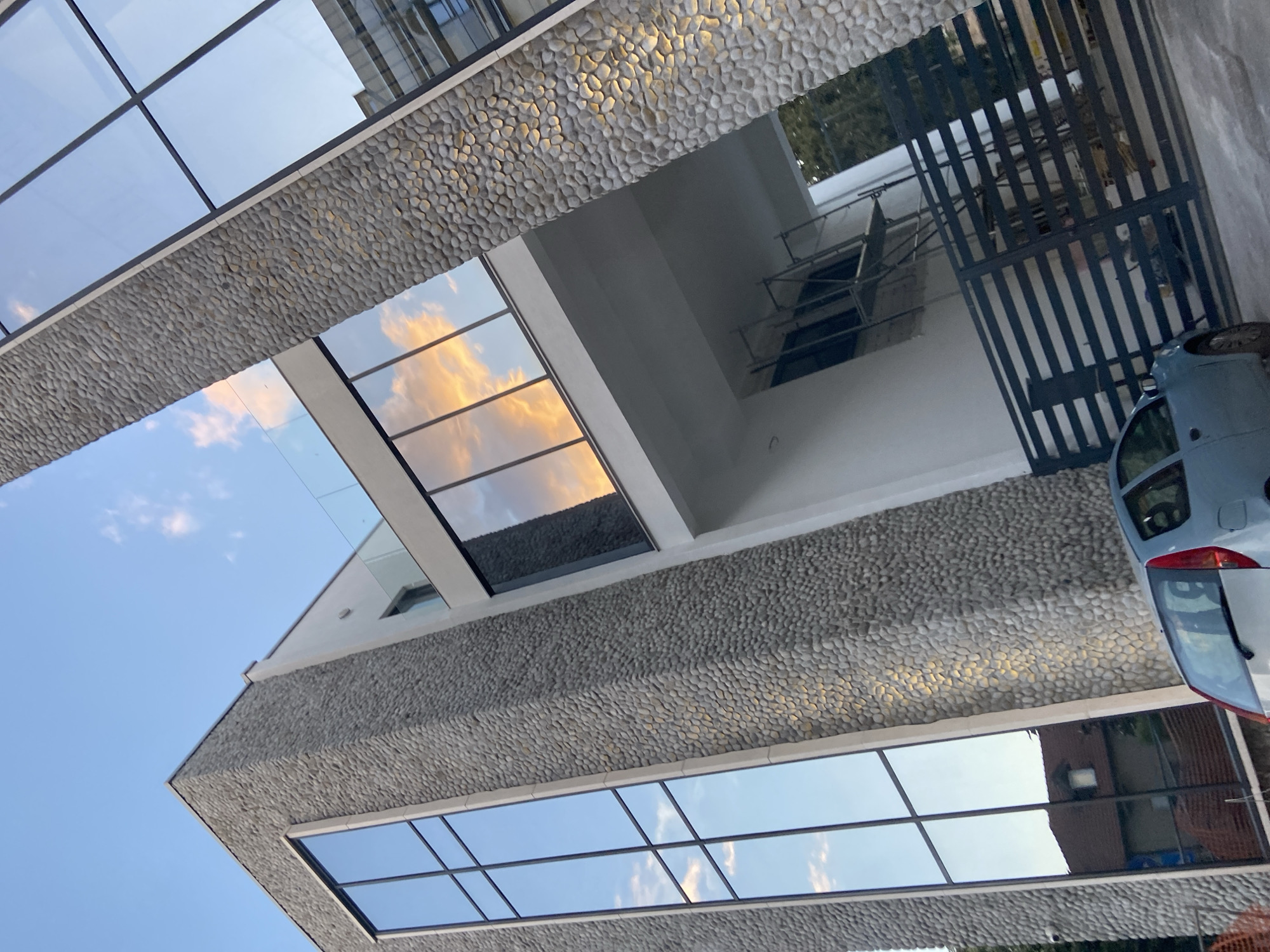 Here is a detailed description of the image:
The image depicts a modern building exterior in Podgorica, Montenegro, with a distinctive design featuring pebble-covered facades flanking large, rectangular window panels. The building's architectural style incorporates clean lines and reflective glass, creating a visual interplay of sky and clouds.
Reflections of the sky, including wisps of clouds, are visible in the windows. The pebble facade creates a textured surface. Scaffolding is visible through a window.
A light blue car is partially visible on the right. A black metal gate is also present. The building appears to be located in an urban area.