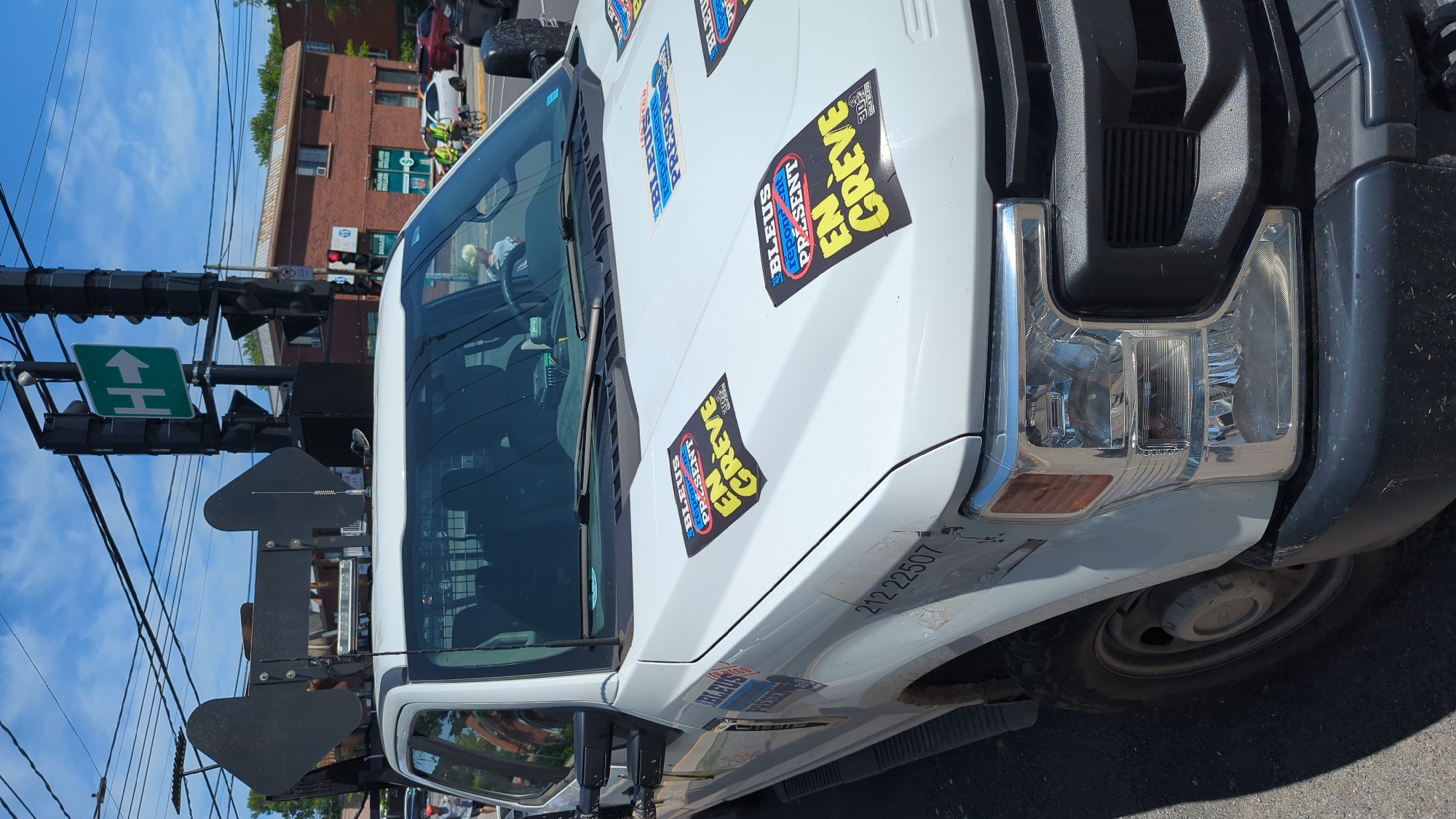 The image shows a white Ford F-series truck parked on a street. The truck is adorned with several stickers that read "BLEUS répondent PRÉSENT en GRÈVE", which translates to "Blues respond PRESENT on STRIKE". These stickers are strategically placed on the hood and the driver's side door of the truck. The truck is equipped with a light bar on its roof. In the background, a traffic light with a green "H" directional arrow is visible, suggesting a possible intersection or parking area. There are also buildings and trees in the background, indicating an urban or suburban setting. The sky is clear and blue with some clouds. The presence of the stickers and the truck's equipment suggest it might be a vehicle used by a unionized group or involved in some form of protest or demonstration. The license plate is not visible, making it difficult to determine the specific region. The context provided by the sticker implies a union dispute, likely within the province of Quebec, Canada, given the French text. The number "212 22507" is faintly visible on the side of the truck.